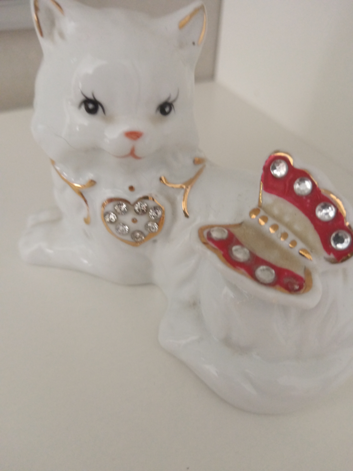 A white ceramic figurine of a cat sits on a white surface. The cat has black eyes, a pink nose, and gold accents on its ears and around its neck. A heart-shaped pendant with clear rhinestones is attached to the cat's chest. A red and white butterfly with gold accents and rhinestones is attached to the cat's back. The background is a plain white wall. The image was taken in Sorochinsk, Russia.