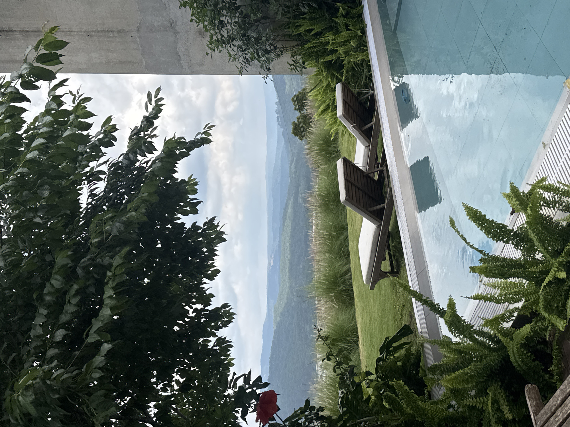 The image depicts a serene poolside scene with a vast, rolling landscape in the background. The foreground is dominated by a swimming pool with crystal-clear blue water, reflecting the sky and surrounding greenery. Two empty lounge chairs are positioned on a neatly manicured lawn adjacent to the pool, inviting relaxation. Lush green ferns and other foliage frame the poolside, adding to the tranquil ambiance. In the distance, a series of mountains creates a breathtaking backdrop, their peaks fading into a hazy sky. The architecture visible on the right side of the image suggests a modern structure, possibly a residence or a resort, built with a light-colored concrete material. The presence of a vibrant red flower on a leafy branch in the upper left corner adds a touch of color to the predominantly green and blue palette. The scene is peaceful and idyllic, suggesting a private getaway in a lush, natural setting. This location is identified as being in Đa Lộc, Vietnam.
