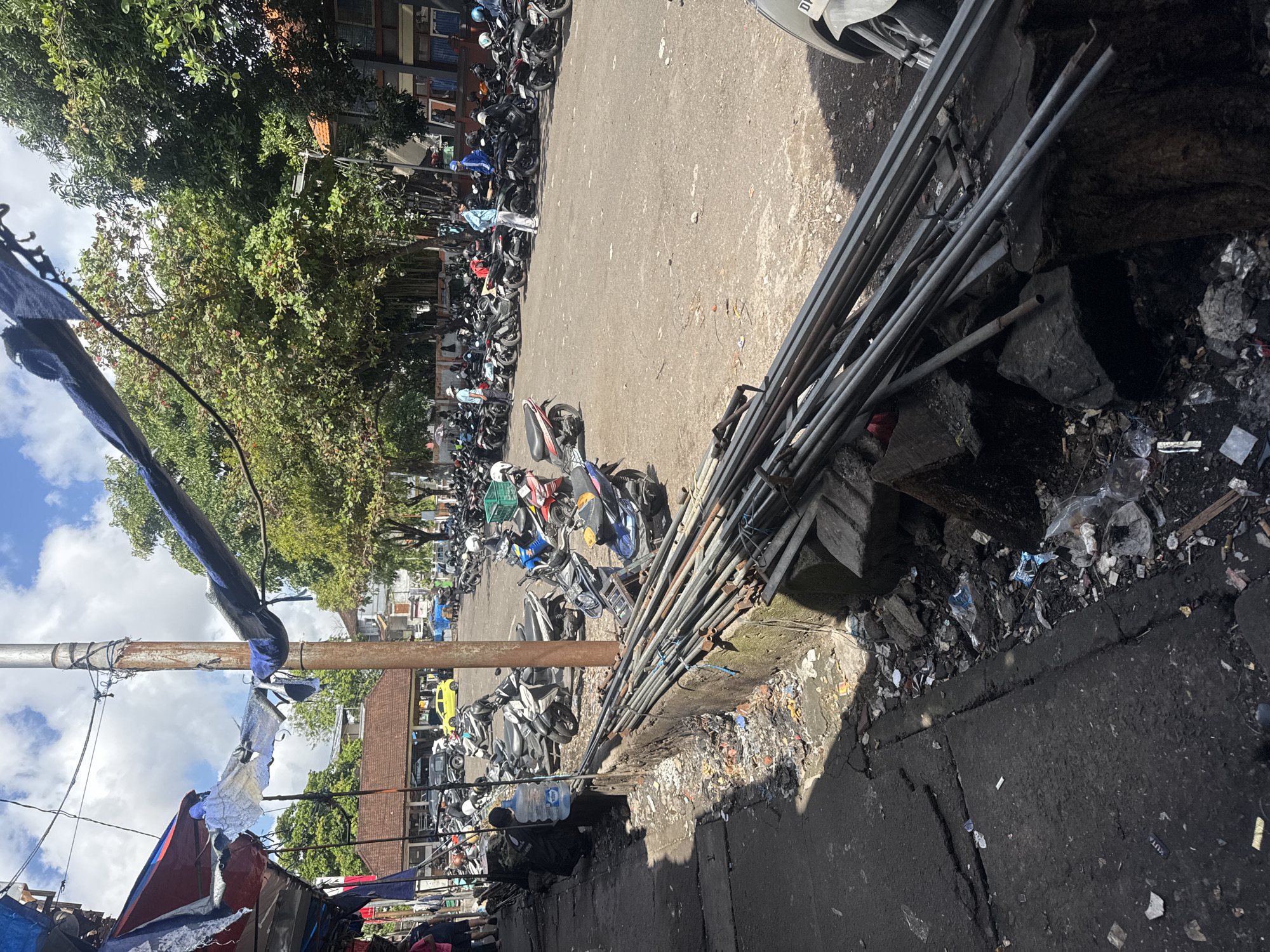 The image depicts an outdoor scene in Denpasar, Indonesia, likely a market or parking area. A significant number of motorcycles are parked on a dirt and asphalt surface. In the foreground, a concrete wall runs along the left side, piled with metallic pipes and debris. The ground next to the wall is littered with trash, including plastic bags, wrappers, and cigarette butts. To the left, a shadowy area suggests a walkway or market stalls with torn tarps hanging from a pole and wires overhead. In the mid-ground, people, some wearing light-colored clothing, are visible near the parked motorcycles, with one individual appearing to carry a large water jug. A bright yellow car is parked further back among the motorcycles. Lush green trees form a canopy in the background, with some buildings visible behind them, one with a distinctive orange-tiled roof. The sky is blue with scattered clouds. The overall impression is of a busy, possibly open-air commercial or transportation hub with evident signs of disarray and litter.