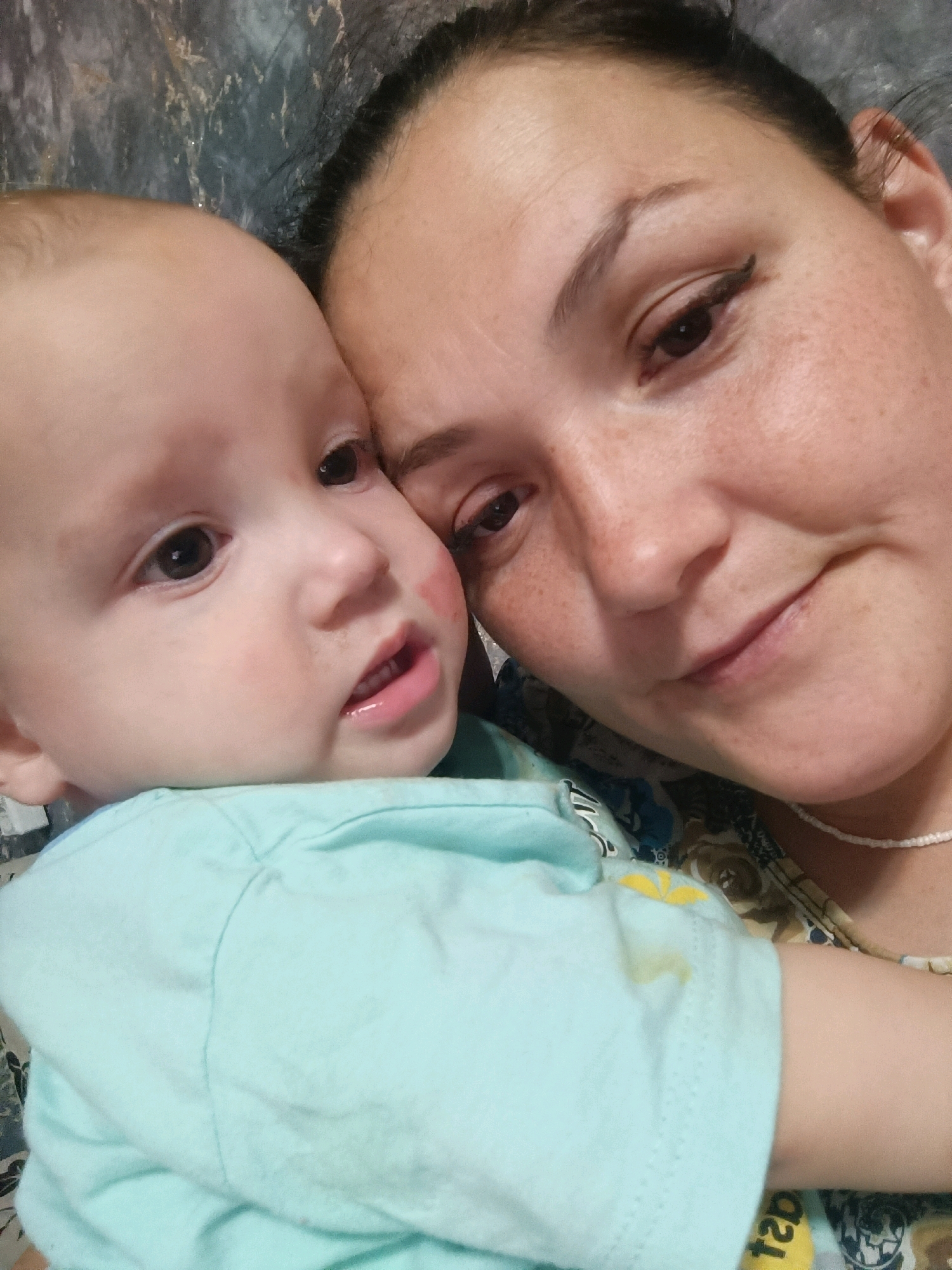 A close-up, overhead selfie features a woman and a baby in what appears to be a domestic indoor setting. The woman, with dark hair and fair skin, is positioned on the right side of the frame, her face angled towards the baby. She is wearing makeup, including winged eyeliner and lipstick. A light-colored beaded necklace is visible around her neck. The baby, on the left, has fair skin and is looking slightly to the right. Red marks are visible on the baby's cheek, possibly from being held or from a rash. Both individuals are wearing light-colored clothing. The background is blurred, showing a patterned wallpaper or fabric, and a dark, textured surface. There is no indication of the city of Karsha, Russia, or any other specific location.