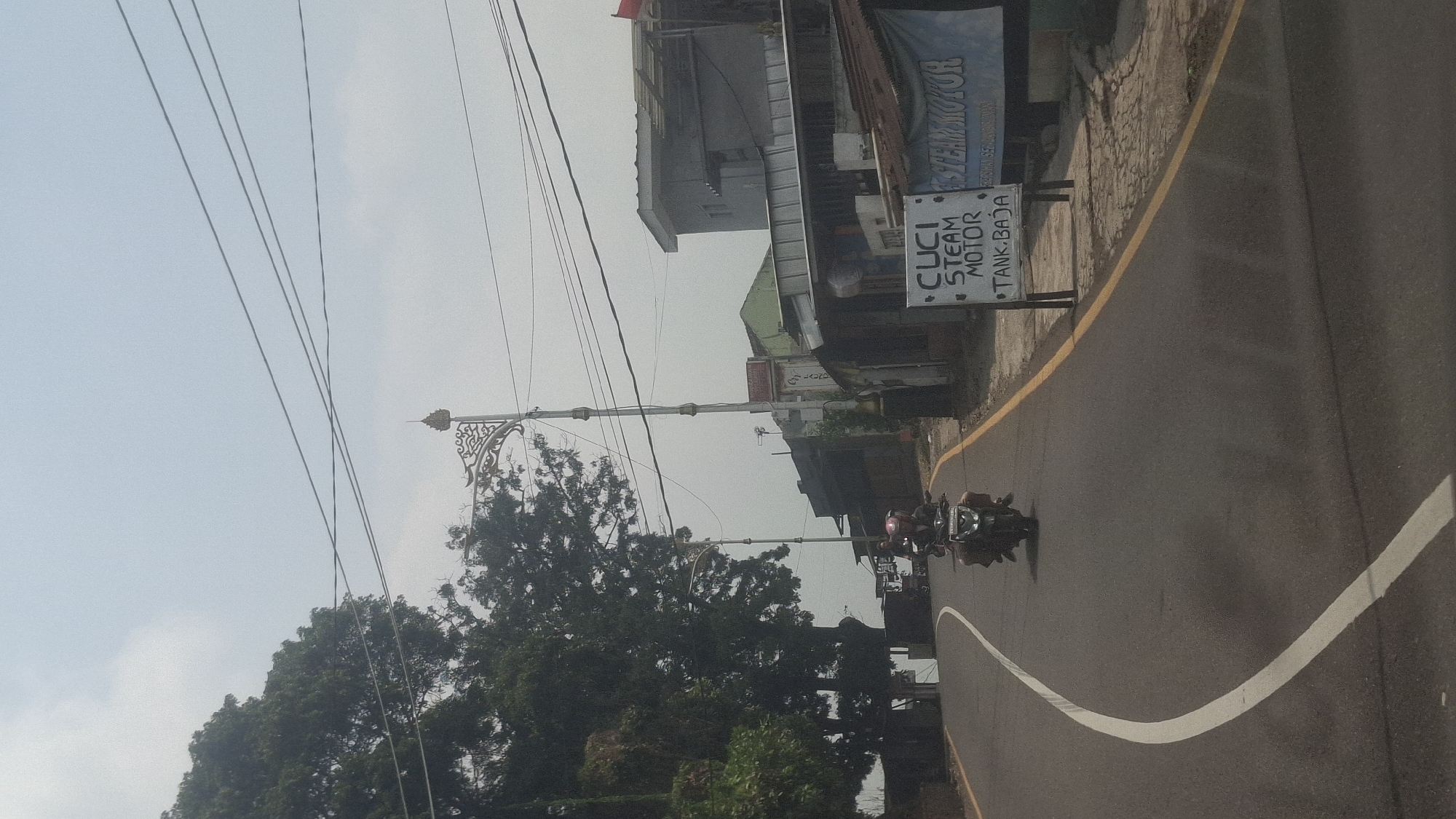 The image depicts a road in Cilaku, Indonesia, under a bright, hazy sky. A paved road curves to the left, marked by a solid white line on the left and a yellow line on the right. A single motorcycle with two riders is traveling away from the viewer on this road. On the right side of the road, there is a small building with a corrugated metal roof and a sign advertising "CUCI STEAM MOTOR TANK. BAJA". Further down the road, more buildings and trees are visible, along with a decorative lamppost. Power lines are strung across the sky, and a large tree with dense foliage is on the left side of the road, obscuring some of the background. The overall impression is that of a rural or suburban setting on a warm day.