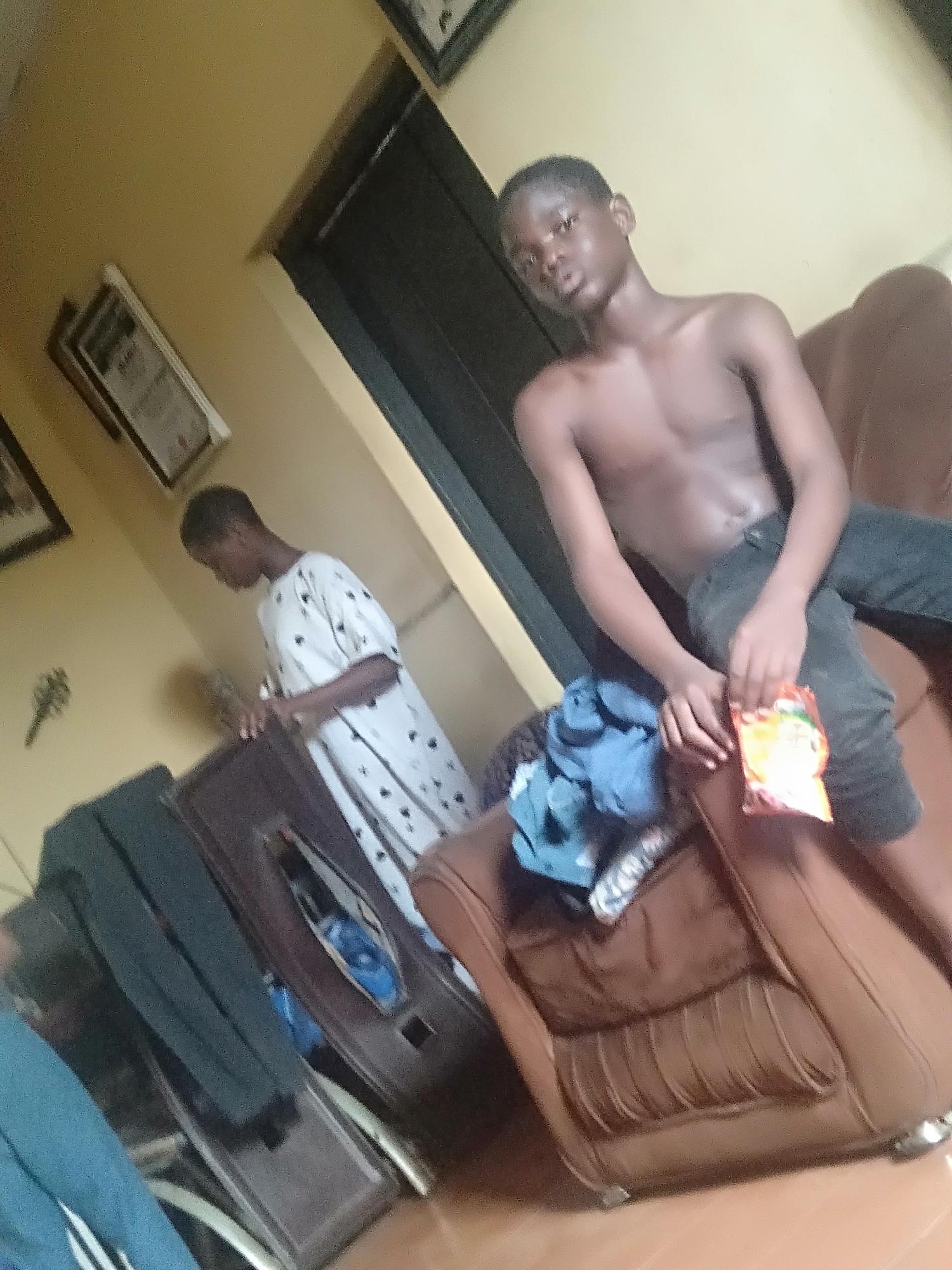 A young, shirtless man is seated on a brown leather armchair in a room, holding a packet of snacks. He wears dark pants. The armchair has a pile of blue clothing on the seat. Behind him, another young person stands at a wooden dresser wearing a white patterned robe. On the wall are framed pictures. The location is likely Shangisha, Nigeria, based on the query.