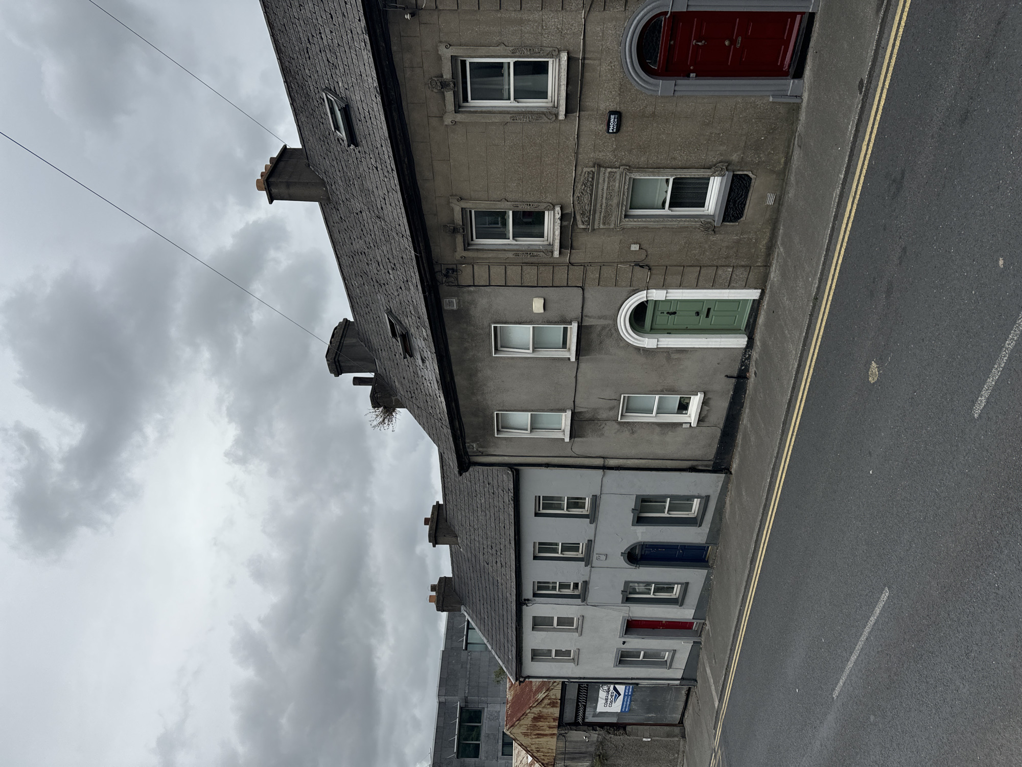 The image shows a row of terraced houses on a street in Waterford, Ireland. The houses are of varying heights and colors, with the closest house on the right being made of grey stone and featuring a large red door. To its left is a narrower house painted a lighter grey, with a green door. Further to the left, the houses are even narrower and painted in lighter shades of grey. The street in front of the houses is asphalt, marked with yellow double lines and a white broken line. The sky is overcast and cloudy. In the background, a modern grey building is visible to the left of the row of houses, along with some industrial-looking structures with rusted metal roofs. The overall impression is of an urban street scene in a historical city.