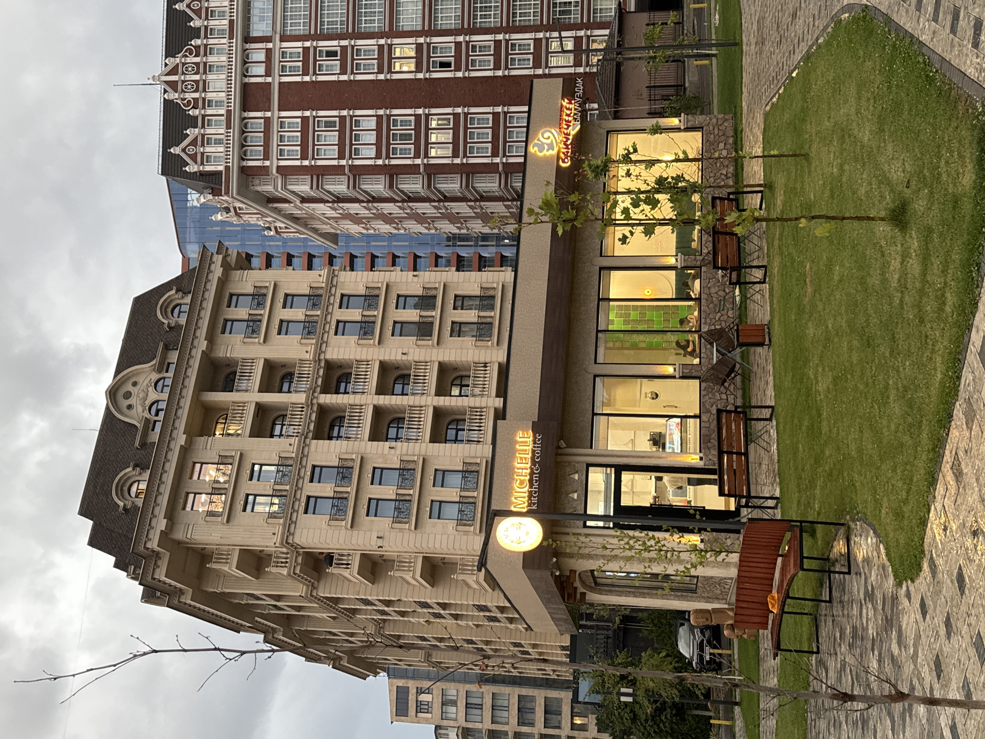 The image depicts a street view in Bishkek, Kyrgyzstan, focusing on a modern cafe with a distinctive architectural style. In the foreground, a paved walkway leads to a grassy area with several young trees. A curved wooden bench is positioned on the left side of the walkway. The cafe, named "MICHELLE kitchen & coffee," features a prominent illuminated sign with the cafe's name and logo. The cafe has large glass windows, revealing an interior with seating and displays. Behind the cafe, a tall, light-colored building with multiple balconies and arched windows dominates the scene. To the right of this building is a taller, red brick building with white decorative trim and numerous rectangular windows. The sky above is overcast. Several cars are visible parked along the street in the background.