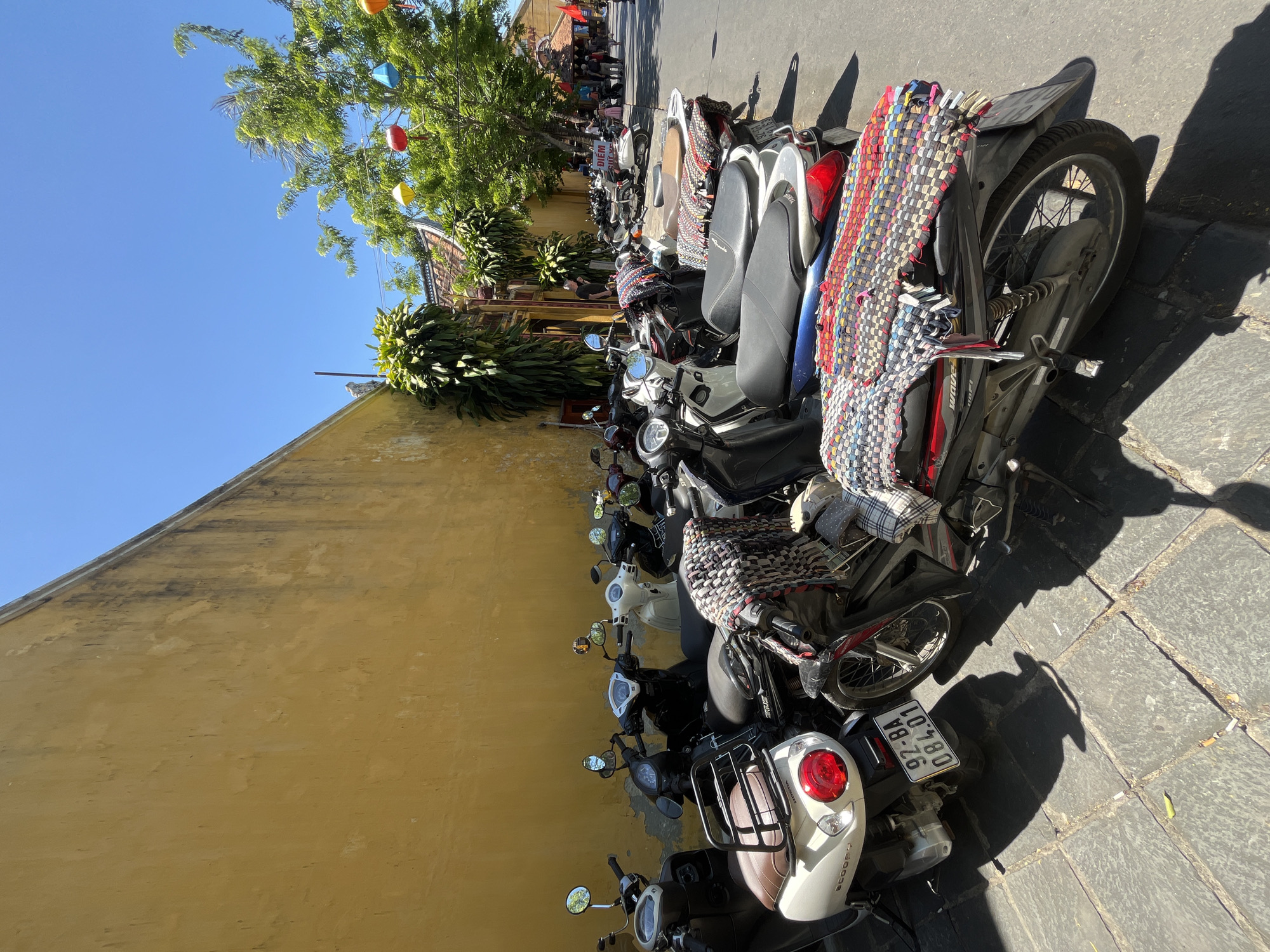 The image presents a vibrant street scene in Hội An Ward, Vietnam, under a clear blue sky on a bright, sunny day. A long row of motorbikes and scooters is parked closely together along a distinctive, textured yellow wall that dominates the left side of the frame.
Lush green trees overhang the scene from the top, adorned with colorful lanterns in red, yellow, and blue, adding to the picturesque ambiance characteristic of Hội An. The ground beneath the vehicles is paved with stone blocks, and the strong, well-defined shadows cast by the motorbikes indicate bright sunlight, likely during mid-morning or early afternoon.
Many of the parked motorbikes feature colorful, woven mats or coverings draped over their seats, a common sight in the region. Visible motorbike models include a "WAVE 100i" and a "SCOOPY". One motorbike's license plate is clearly legible as "92-BA 084.01". In the background, beyond the dense line of motorbikes, glimpses of traditional Vietnamese architecture and a few people can be seen further down the street. A partially visible sign on a distant building appears to read "DIEM SUE", potentially indicating a shop. The scene conveys the bustling yet charming atmosphere of a historic Vietnamese town.