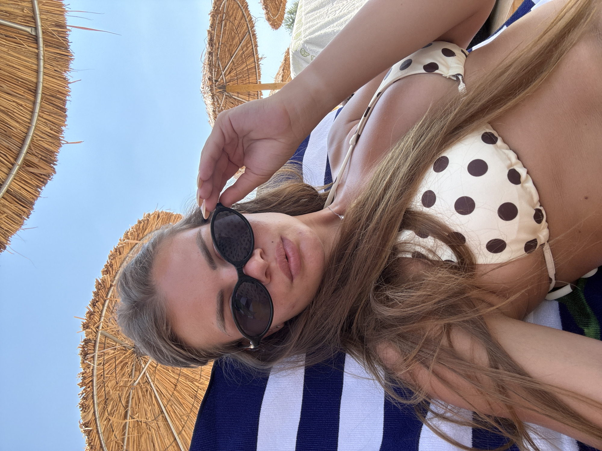 A young woman with long, light brown hair is lying on a beach chair, wearing a polka dot bikini and sunglasses. She is resting under a thatched umbrella on Ofrynio Beach, Greece, with the blue sky visible above. Her right hand is resting on the side of her sunglasses. The beach chair is covered with a striped towel.