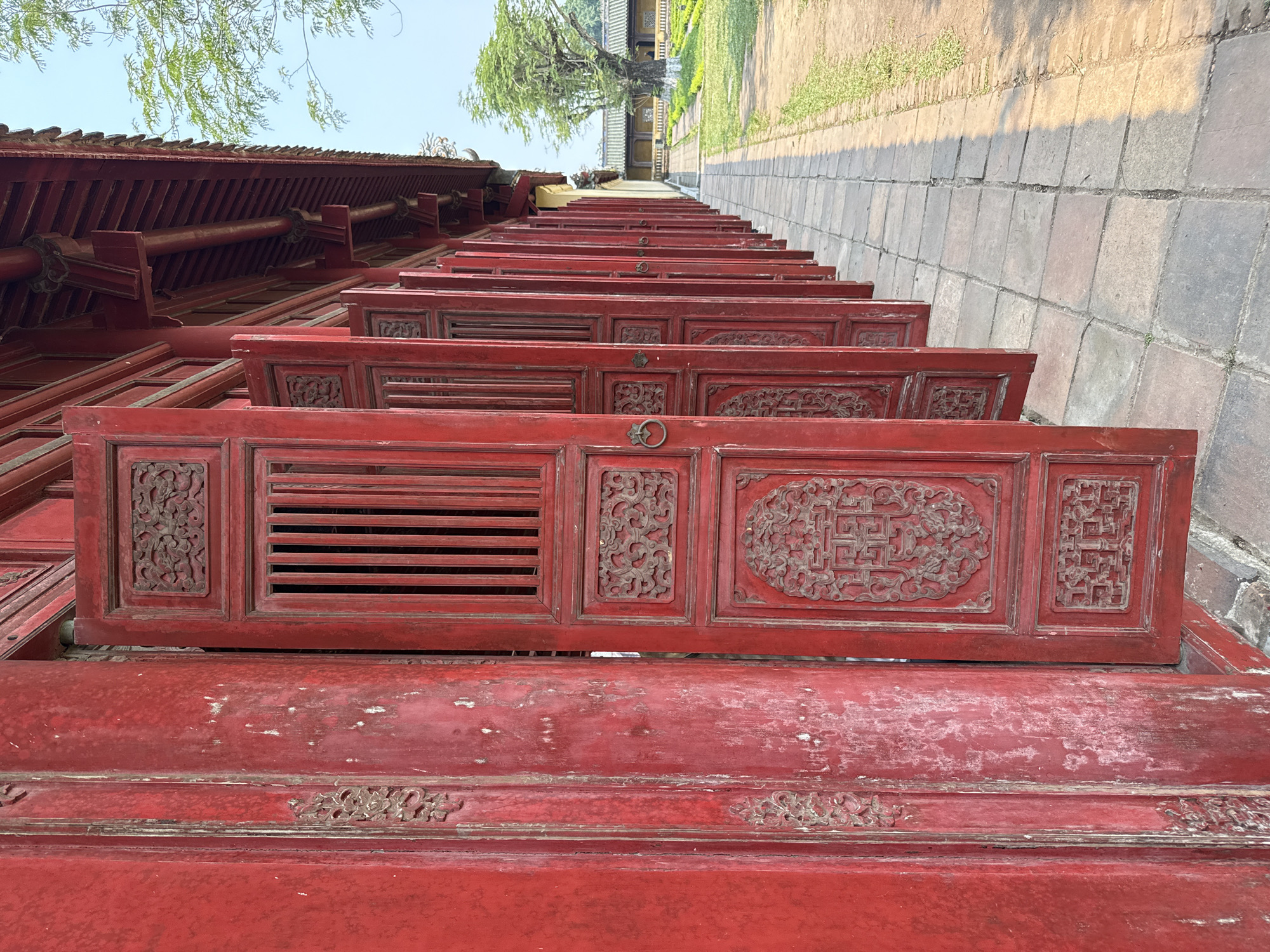 The image shows a row of traditional red Vietnamese doors, possibly from a historic structure, lined up against a stone wall or walkway. The doors are adorned with intricate carvings and designs, some featuring lattice-like ventilation and others ornamental motifs. Their weathered appearance suggests age and exposure. The location is Phú Xuân Ward, Vietnam, identifiable through the style of architecture.
