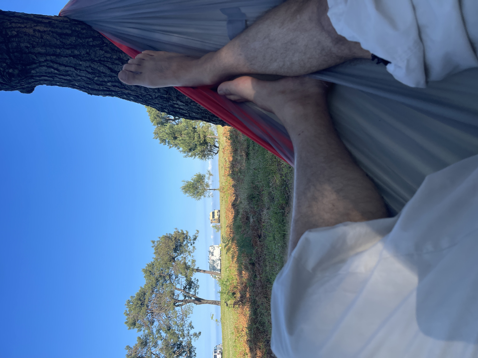 The image shows a person lying in a hammock tied to a tree. The hammock is red and gray. The person's legs and feet are visible, covered in light-colored pants. The hammock is positioned in a grassy area with several trees in the background. In the distance, there are a few RVs parked on a hill, suggesting a camping or recreational setting. The sky is clear and blue. The overall scene is peaceful and outdoorsy. This location is not identified as being in Kobuleti, Georgia.