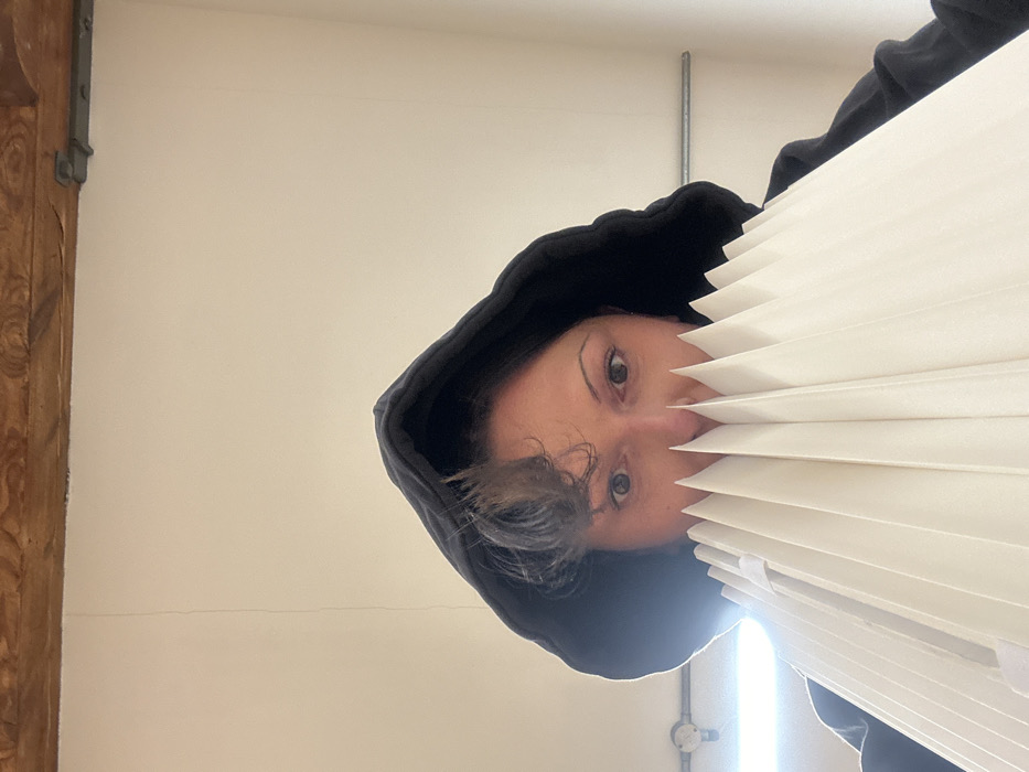 A person wearing a black hooded sweatshirt looks at the camera from below while holding a white, pleated, fan-like object that covers most of their face. Their eyes, nose, and a small section of gray hair are visible. The background consists of a white wall, a wooden doorframe, and a silver pipe. The image appears to be taken from a low angle and then rotated counterclockwise.