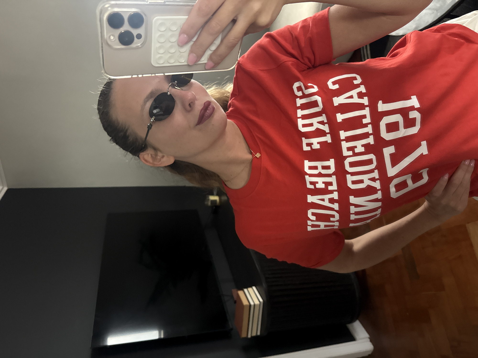 A woman is taking a selfie indoors. She is wearing a red t-shirt with white lettering that reads "California, Surf Beach, 1952." She is also wearing small, round sunglasses. The woman has light skin and is wearing lipstick. She is holding a phone with a clear case that has a white, textured grip. In the background, there is a dark gray wall with a television, a stack of books and a portion of wood flooring visible. The location of Cape Town, South Africa cannot be determined from the image.