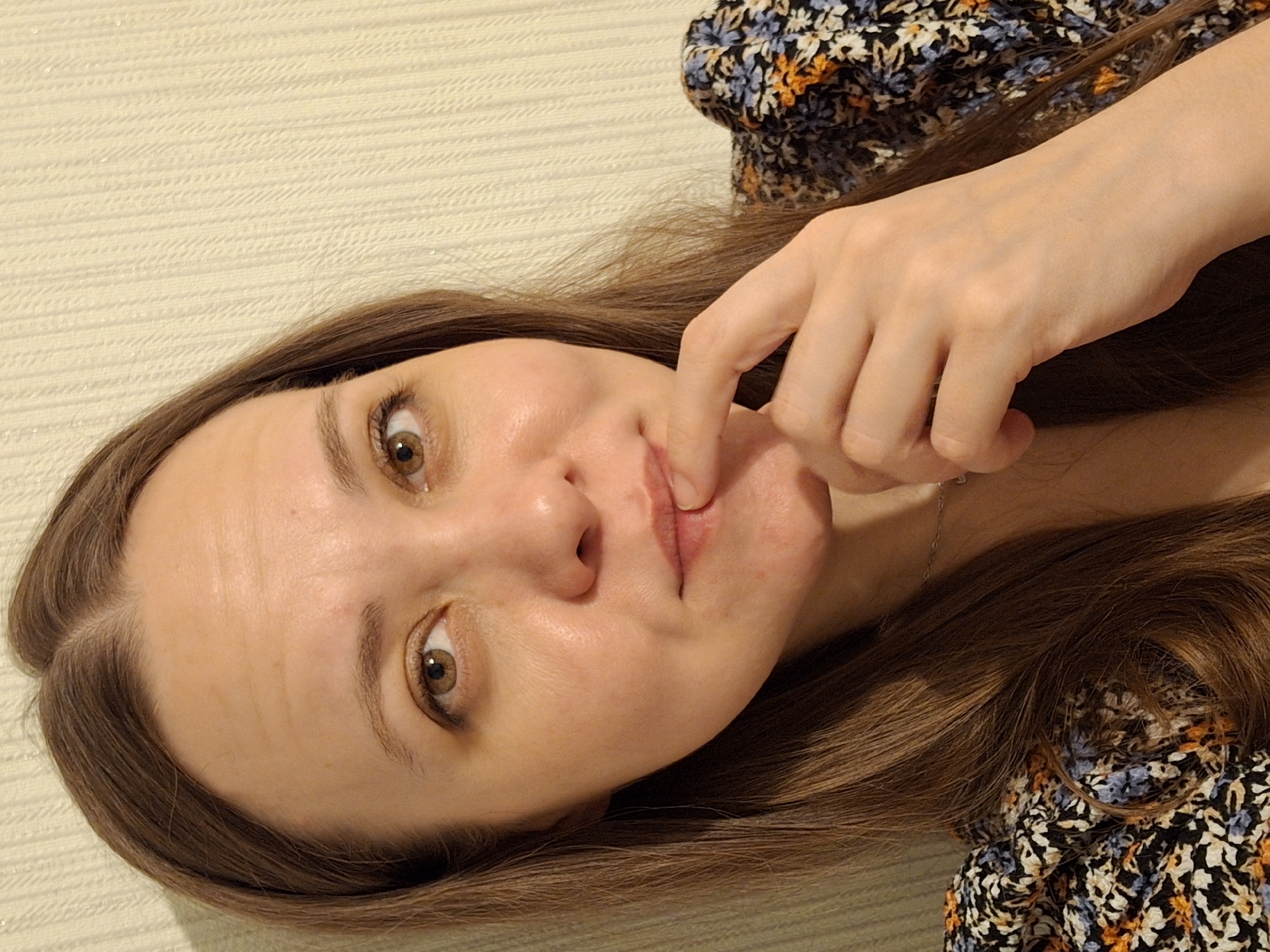 A woman with light skin, brown hair, and hazel eyes is lying down, her head resting on a light-colored textured surface. She is wearing a floral-patterned top with black, white, blue, and orange flowers. Her right hand is near her mouth, with a finger touching her lower lip. A thin silver chain is visible around her neck. The scene is indoors with no other prominent environmental details to suggest the location in Unknown, Unknown.