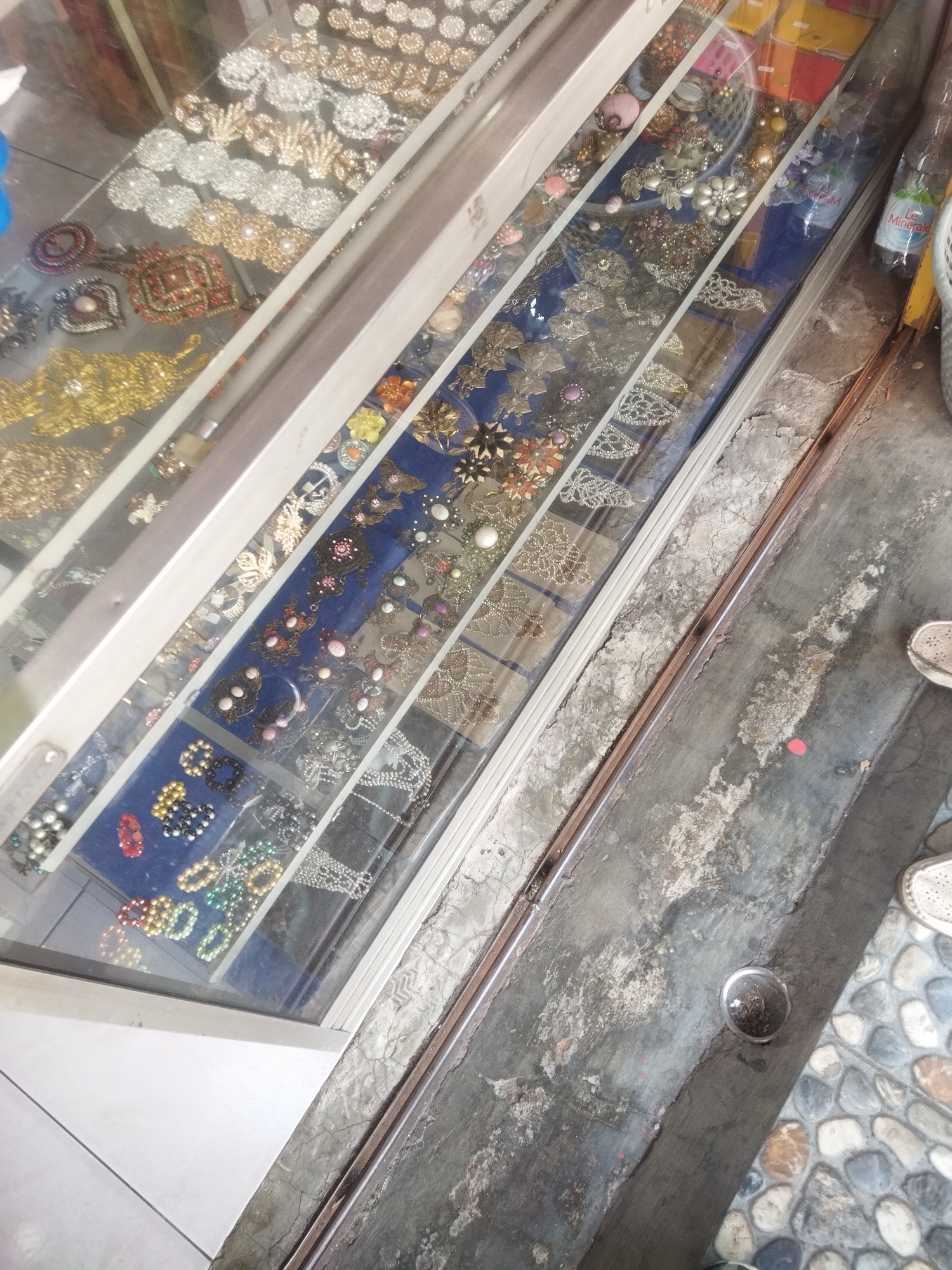The image shows a display case filled with various accessories, possibly for sale at a shop. The display case is made of glass and metal, and it is filled with rows of jewelry and decorative items. The items include necklaces, bracelets, earrings, and brooches, many of which are adorned with beads, rhinestones, and intricate metalwork. Some of the items are gold-colored, while others are silver or have colorful embellishments.
The display case is placed on a surface that appears to be a floor made of concrete and tiles. In the background, there are glimpses of what could be shelves or other merchandise. There is also a bottle of mineral water on a shelf to the right of the display case, indicating it is likely a retail environment.
The scene appears to be an indoor setting, possibly a small shop or market stall. The lighting is somewhat dim, but the items in the display case are clearly visible. The overall impression is one of a place selling a variety of small, decorative items.
The location is identified as a shop in the city of Wonogiri, Indonesia. This specific detail suggests the image was taken in that particular geographic area. Without further information or visual cues in the image itself, it is difficult to ascertain the exact type of shop or the specific street or area within Wonogiri. However, the presence of retail items and a display case strongly points to a commercial establishment.