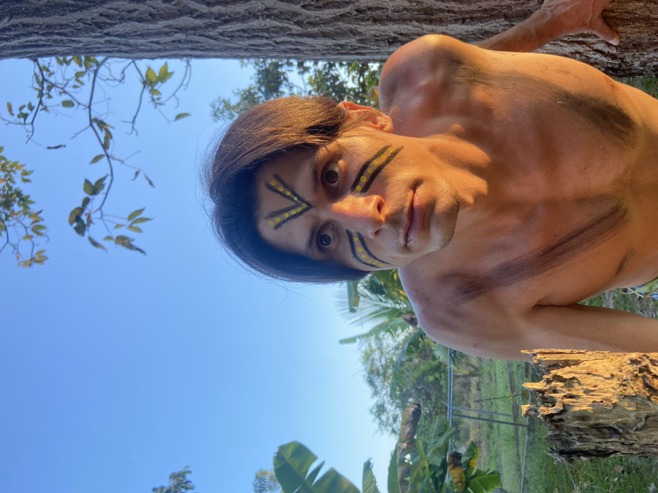 The image depicts a male individual positioned outdoors in Los Velásquez, Venezuela. He has medium-length dark hair and is shirtless, displaying body paint. The facial paint consists of black 'V' shaped lines with yellow dots on the forehead, and two horizontal black lines with yellow dots under each eye. Black diagonal lines are visible across his upper chest and shoulders. He is in a crouching stance, with his right hand resting on a tree trunk to his right. His brown eyes are directed upward and slightly to the right. The scene is illuminated by warm, direct sunlight under a clear blue sky. The background features green banana plants with large leaves on the left, additional green foliage, and other trees. A faint metal fence structure is discernible in the mid-ground. In the lower-left foreground, a weathered tree stump is prominently lit. The ground is covered with green vegetation.