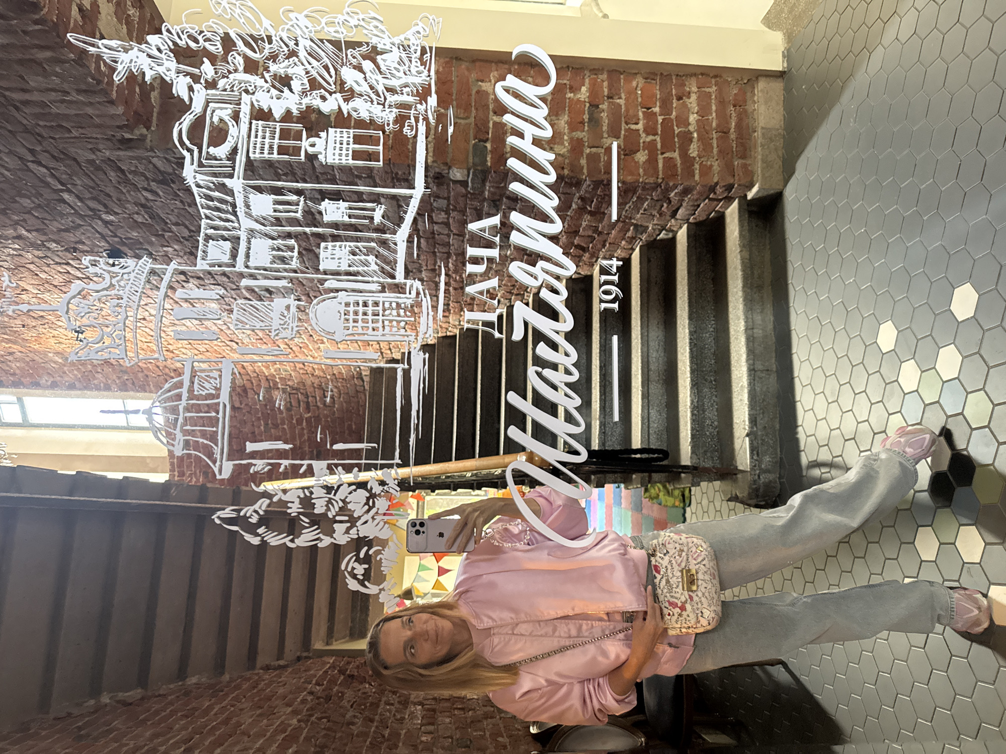 A woman is taking a selfie in a mirror. She is wearing a light pink bomber jacket, grey jeans, and pink sneakers. She is carrying a small, patterned crossbody bag. The background is a brick wall with a staircase leading up. White line art of buildings and decorative elements are visible on the mirror. Text on the mirror reads "НАЧАЛО" and "1914". The lighting suggests it is daytime, possibly indoors. The woman's expression is neutral. The setting appears to be a historic building in Saint Petersburg, Russia, perhaps a cafe or a museum with architectural decorations.