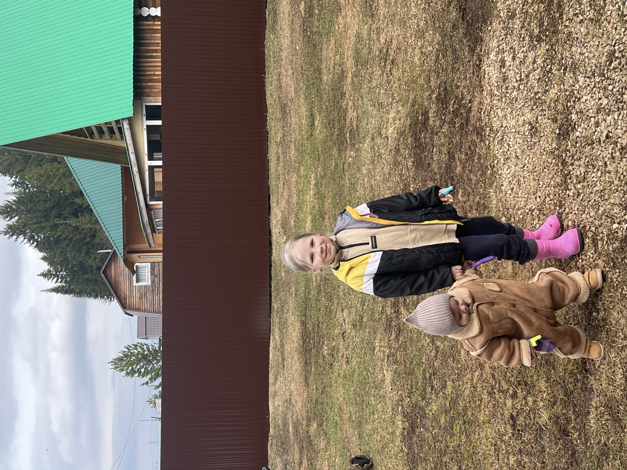 Two young children are in a grassy yard. One child, wearing a black, yellow, and white jacket, dark pants, and pink boots, is sitting. The other child, dressed in a brown jumpsuit with a grey hat, is lying down. They are both holding small, colored objects. Behind them is a brown fence, a wooden building with a green roof, and trees. The ground is a mix of grass and gravel. The scene is likely in Protasy, Russia, indicated by the roof-type on the main structure.