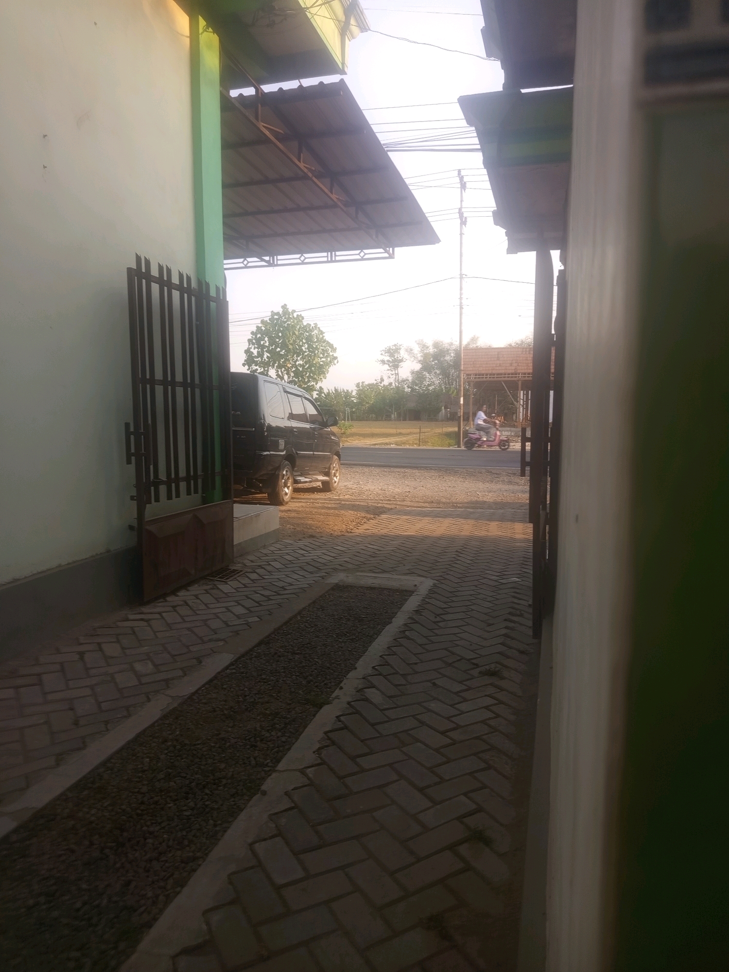 In Cangakan, Indonesia, a paved walkway with a drainage channel filled with small stones runs alongside a building with a light-colored wall and a green vertical beam. A dark metal gate, partially open, stands on the left. Beyond the walkway, a dark-colored van is parked in front of a raised concrete area. Further in the background, a paved road is visible, with a person riding a pink scooter. The environment features scattered trees and a clear sky.
