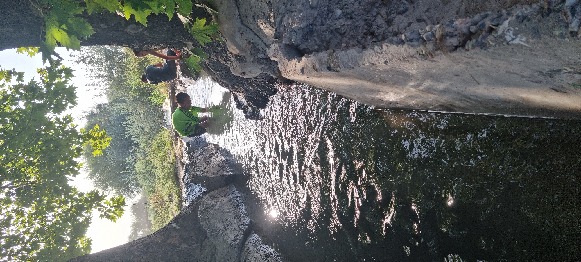 The image depicts a canal with a young boy in a green shirt and shorts wading in the water, reaching down. To his right, two other young individuals are seated on the canal's edge, partially obscured by a tree. The canal is surrounded by rough, rocky terrain and dense foliage. Sunlight glints off the water's surface, creating shimmering reflections. The scene appears to be in an outdoor, natural setting, possibly a rural area or park within the city of Ҷомӣ, Tajikistan, given the context of the question.