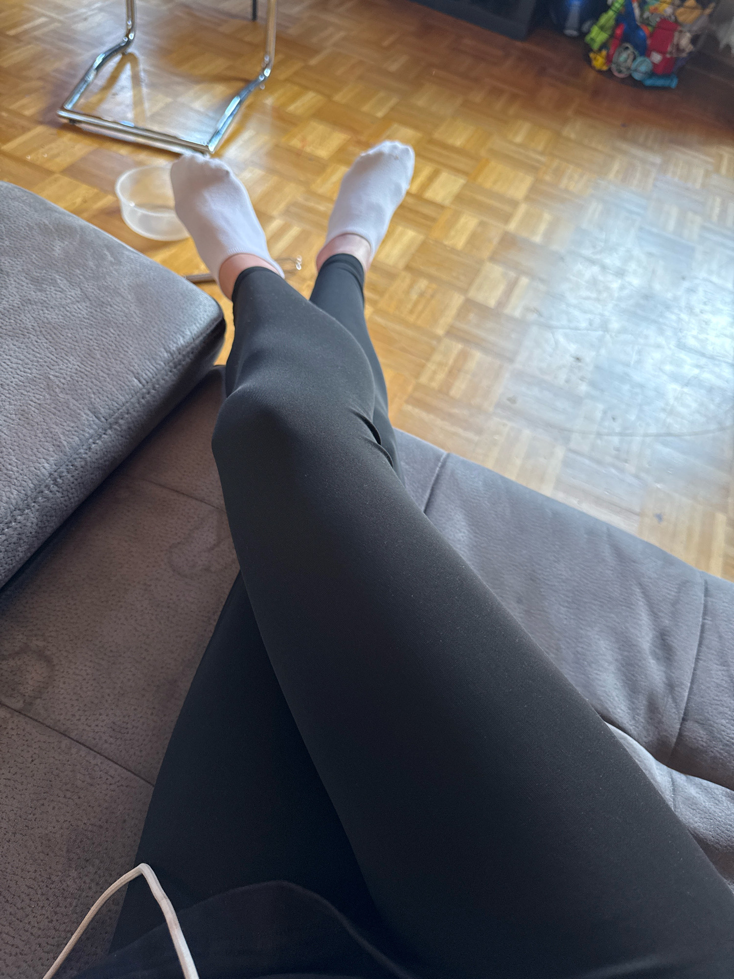 The image displays a close-up view of a person's legs, from the knees down, resting on a dark gray sofa. The person is wearing white ankle socks and black leggings. Their legs are crossed at the knees. The background shows a wooden parquet floor and a portion of a metal chair with a transparent bowl underneath it. A basket with various colorful objects, possibly toys, is partially visible in the upper right corner. The lighting suggests an indoor setting. The location in the city of Überherrn, Germany, is not identifiable from the visual information provided in the image.