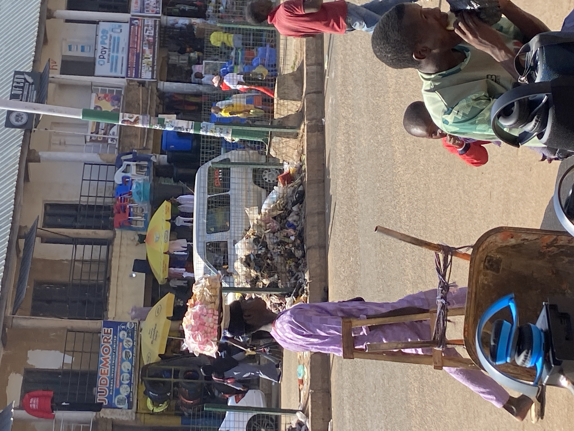 The image depicts a street scene in Bukuru, Nigeria. A man is seated on a rudimentary wooden chair, possibly stationary, positioned in the street. He is wearing a light purple shirt and carrying a headload wrapped in pink and white material, appearing to be selling something. Behind the man, there's a parked white van and a significant pile of trash along the curb. Across the street are storefronts with signs including "JUDEMORE" and "PayPOS". Two children are present, one eating and the other possibly watching. Another man wearing a red shirt is standing on the opposite side of the street. Other individuals are visible in the background, near the storefronts.