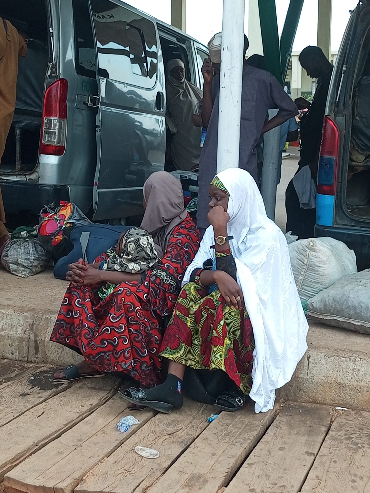 The image shows an outdoor scene near a vehicle, possibly a transport hub, in Gombe, Nigeria. Two women are seated on a concrete ledge with wooden planks in the foreground. The woman on the left wears a red and black patterned dress and a brown hijab. She is leaning forward with her hands clasped. The woman on the right wears a white hijab and a colorful patterned dress with yellow, red, and green designs. She is looking to the left, with her hand resting on her chin. Behind them, a grey van with its doors open is visible. Several large bags, possibly luggage, are placed near the van. Other people are present in the background, some standing near the van. The environment appears to be a dusty, open area, likely a bus station or market.