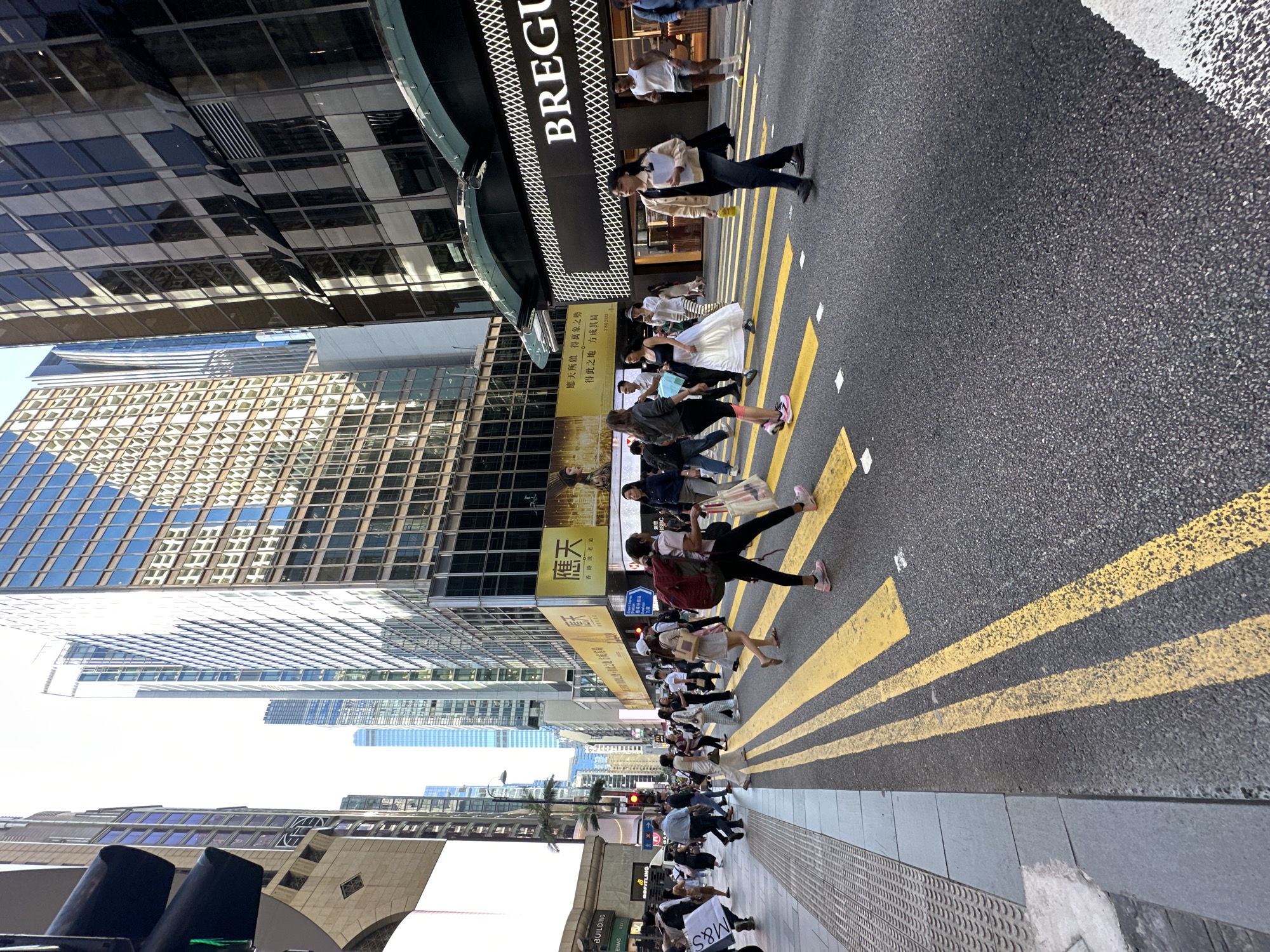 The image shows a busy street scene in Hong Kong, China. The photograph is taken from a low angle, looking across a crosswalk at a wide, multi-lane street lined with tall buildings. Yellow crosswalk lines are painted on the asphalt road. Several pedestrians are crossing the street, some carrying shopping bags. On the right side of the street, a large, illuminated sign for "BREGUET" is visible, indicating a luxury goods store. The buildings are modern, with many windows reflecting the sky and surrounding structures. A traffic light is visible on the left side of the image. The overall atmosphere is one of a bustling urban environment during daytime.