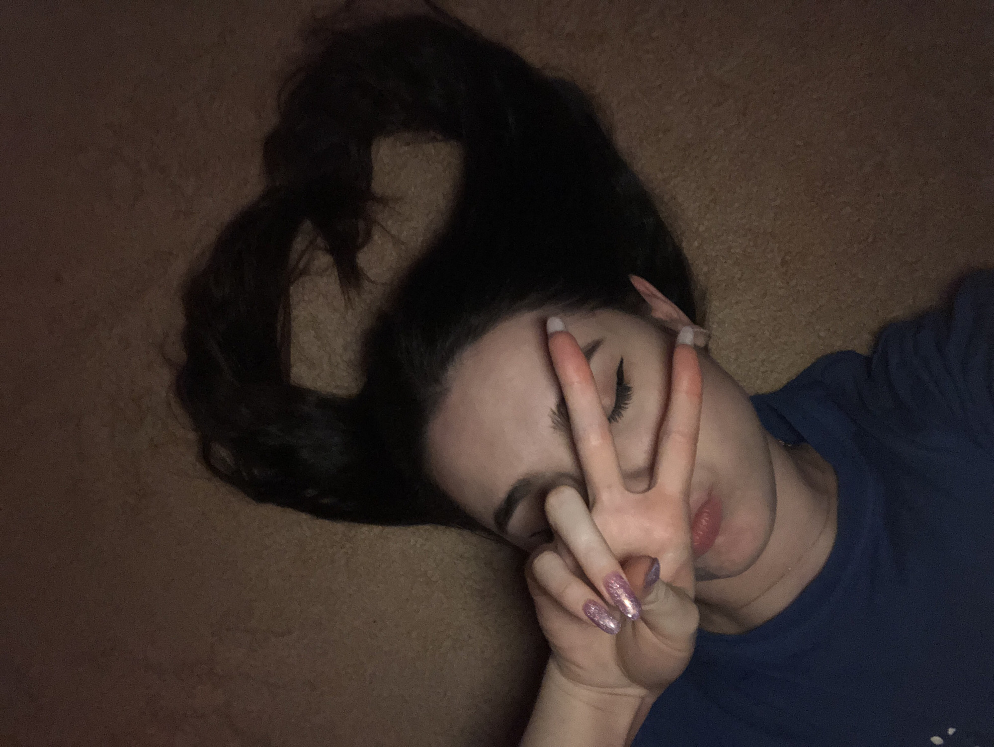 A young woman is lying on her back on a tan carpet. Her dark hair is spread out around her head, with a portion of it styled to resemble a heart shape. She is holding up her hand in a peace sign, partially covering her face. Her nails are long and glittery. She is wearing a dark blue t-shirt. The lighting in the room is dim. The location is unknown, as no information in the image indicates a location in the city of Noviy, Russia.