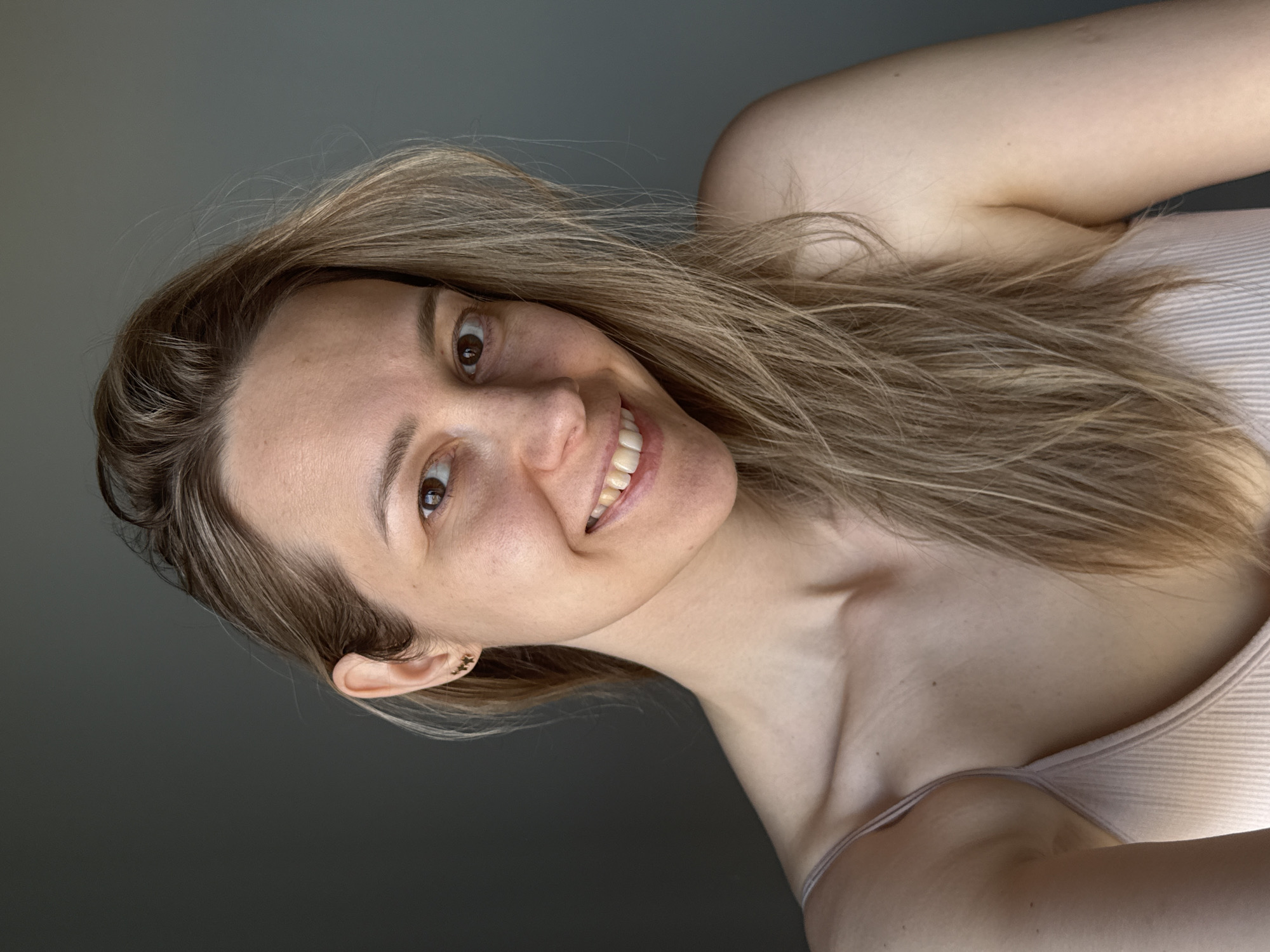 A woman with long blonde hair and brown eyes is smiling at the camera. She is wearing a light pink tank top. She has a fair complexion and a gold stud earring in her left ear. The background is a solid grey color. This image does not contain information to indicate that this was taken in Chaikovsky, Russia.