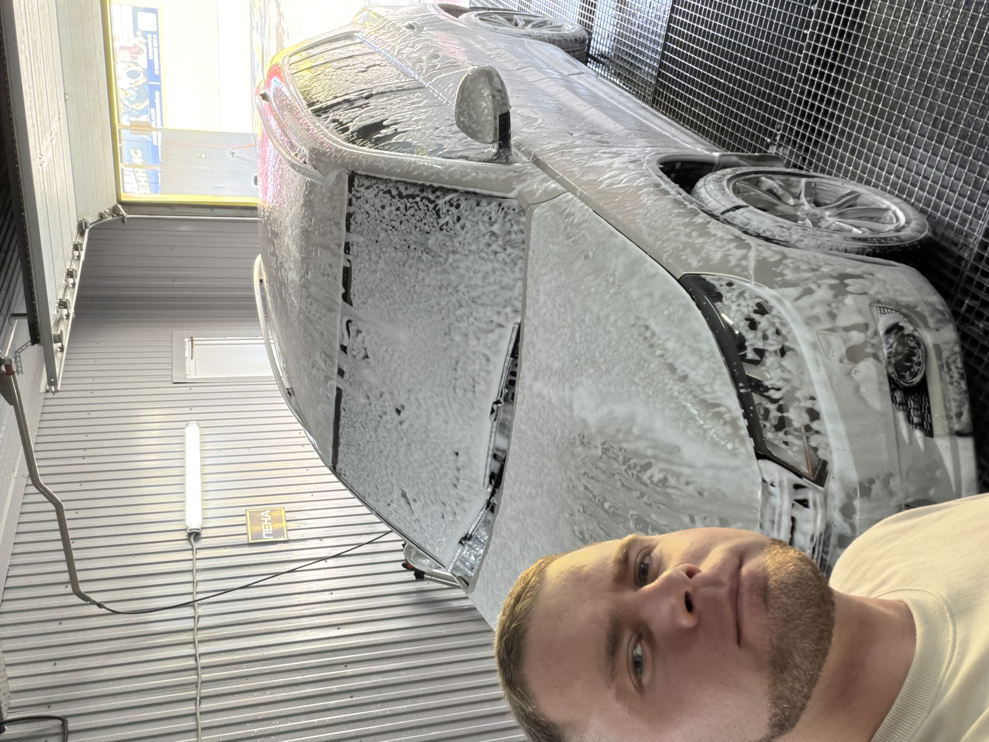A man with a short, light-brown haircut and a beard is taking a selfie in what appears to be a car wash in Chaikovsky, Russia. The car is covered in soapy foam. The car wash has metal walls and ceilings. A long fluorescent light is mounted on the wall along with a sign with "ПЕНА" (foam) written on it. The car wash door is open, letting in daylight.