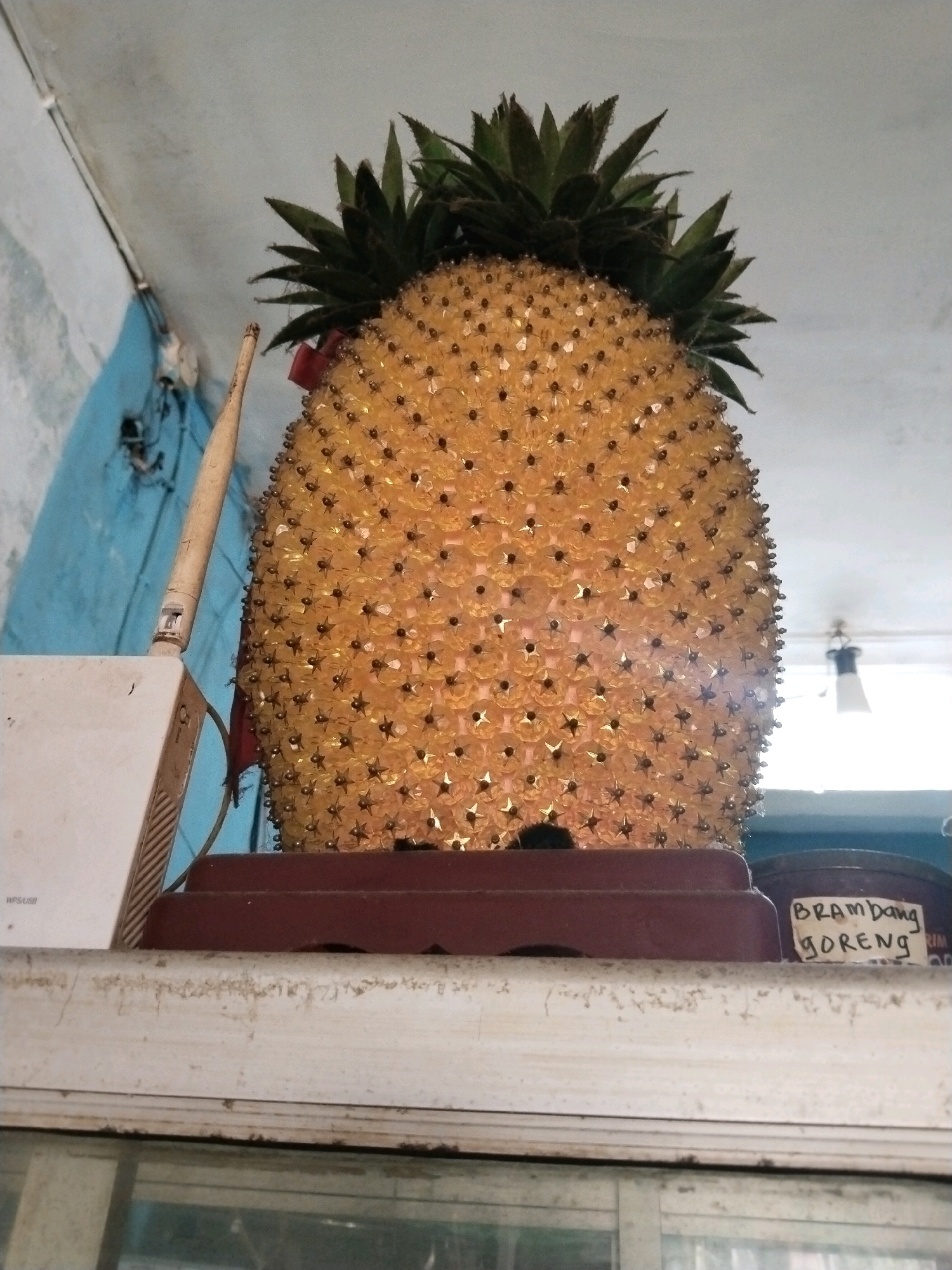 The image shows a decorative pineapple ornament made of yellow plastic star-shaped pieces and brown beads, placed on a shelf. To the left of the pineapple is a white Wi-Fi router with an antenna. To the right, a brown container with a handwritten label that reads "BRAMBANG GORENG" is partially visible. The background consists of a blue wall and a white ceiling with a light fixture. The setting appears to be a room in a building in the city of Prigi, Indonesia, possibly a home or a small shop.