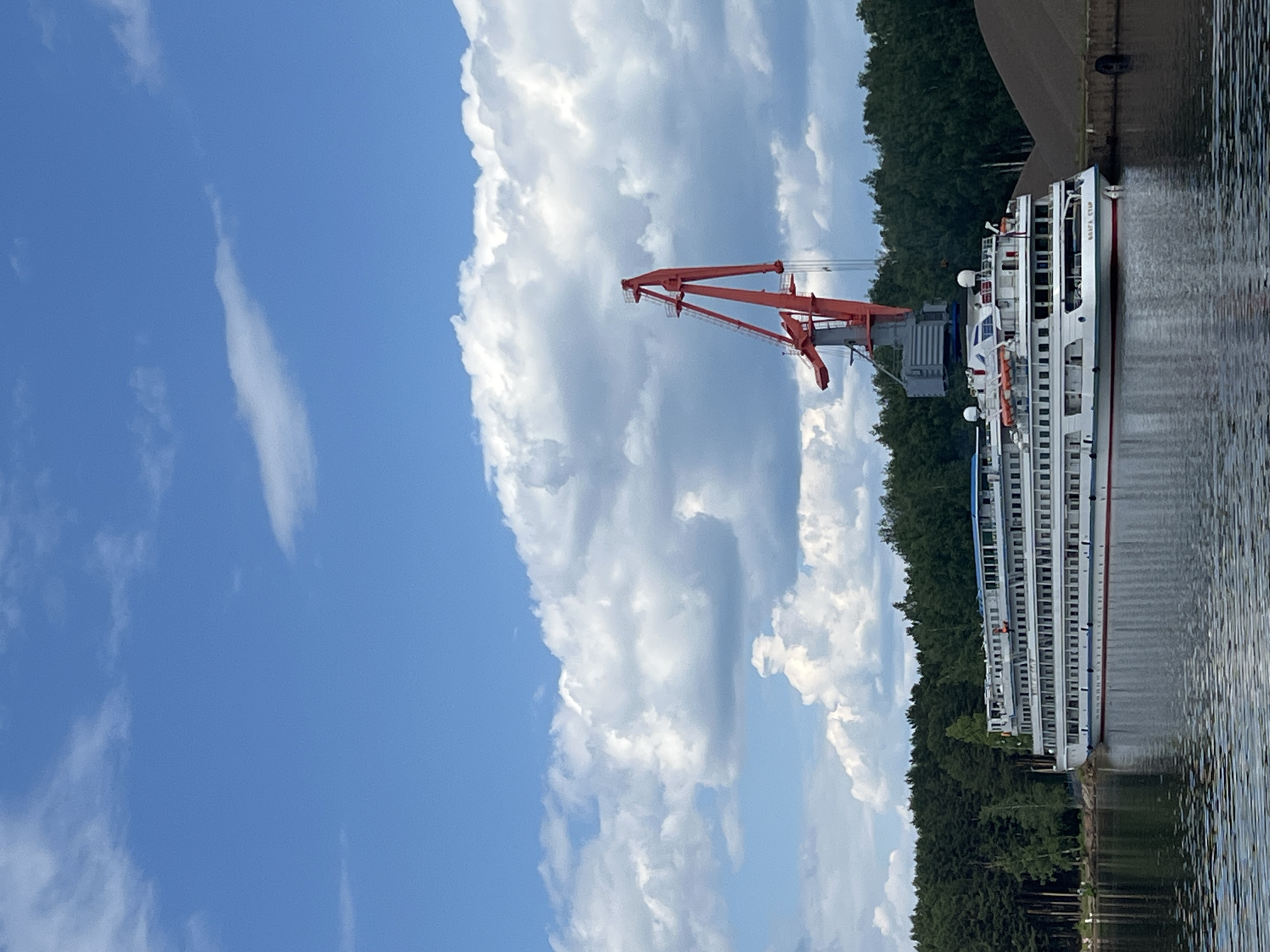 A large white cruise ship is docked in a body of water, likely a river or canal, with a dense green forest lining the background. A tall red gantry crane stands behind the ship, towering over it. The sky is a vibrant blue with scattered white clouds. The ship is multi-decked and appears to be a passenger vessel. To the right of the ship, a large pile of dark material, possibly coal or gravel, is visible next to a concrete structure. The water is calm, reflecting the ship and the surrounding trees. The scene is likely a port or loading area in the city of Chaikovsky, Russia, given the presence of the crane and the cruise ship.
