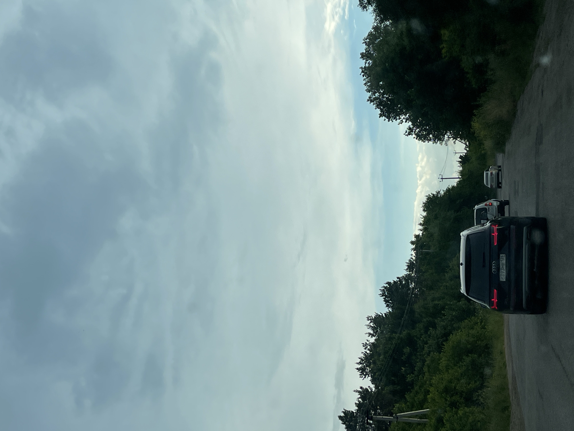 An overcast sky looms above a paved road with a dark Audi SUV in the foreground, its taillights illuminated. The SUV is in the right lane of a two-lane road, with another car visible further ahead in the same lane. To the left of the road, a wooden utility pole and dense green trees line the median. On the right side of the road, more lush green trees and vegetation form a dense embankment. The road appears to be in a rural or semi-rural area, possibly on the outskirts of Tatarqa, Russia, given the presence of utility poles and the natural landscape. The overall impression is a daytime scene, likely in the summer or late spring, with a hint of impending rain due to the cloud cover.