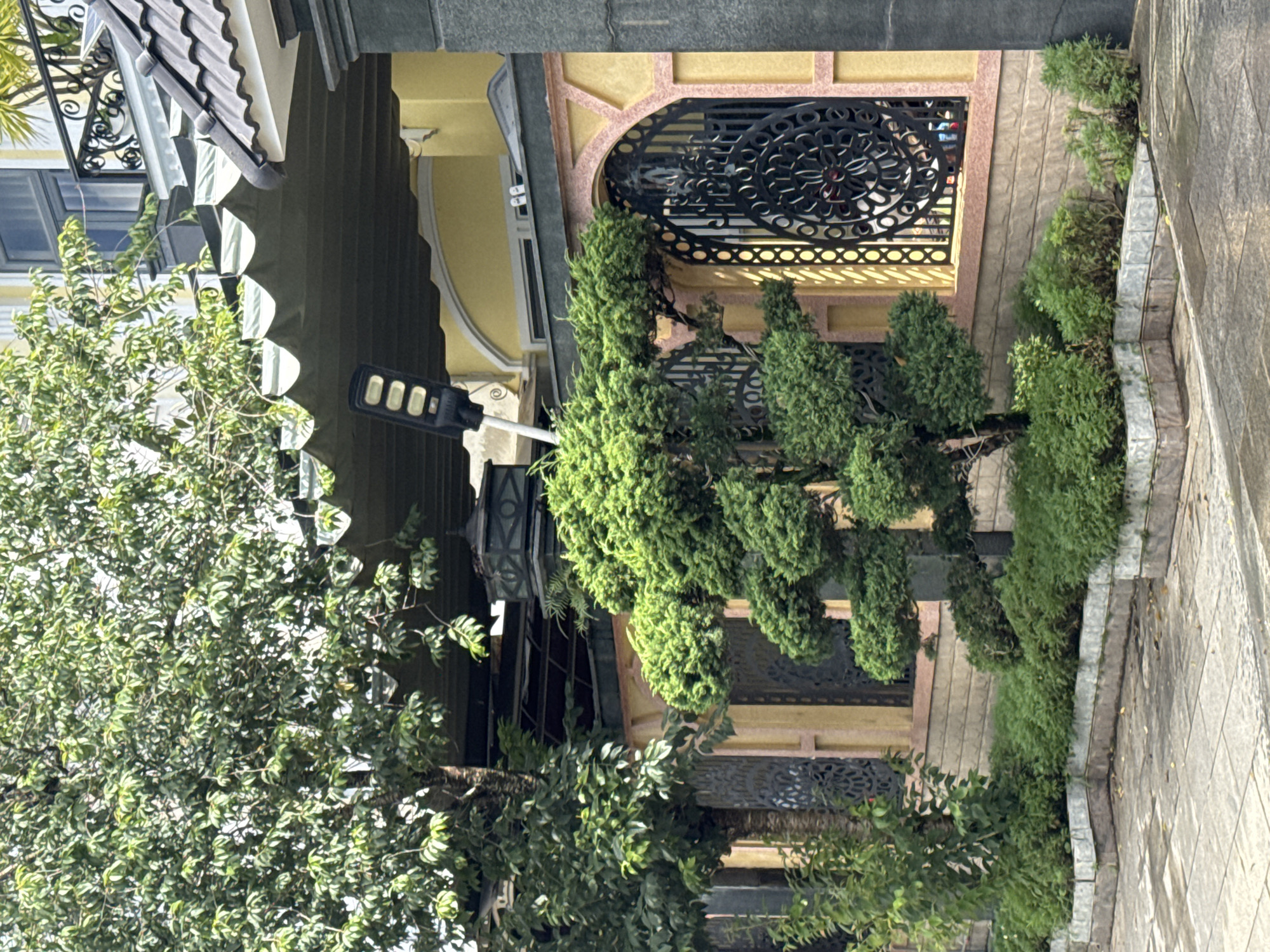 In the city of Lâm Viên - Đà Lạt, Vietnam, a well-maintained yard features a variety of plants and architectural elements. A large tree with dense green foliage dominates the upper left portion of the frame, its branches extending over a grey, corrugated awning. To the right, a black solar-powered street lamp with three LED panels stands beside the awning. Beneath the awning and tree, a decorative stone wall with an arched window framed by ornate black metalwork is visible. The wall is painted yellow with pink accents. In front of the wall, a tiered garden bed is filled with lush green shrubs. A sculpted evergreen tree, shaped in a tiered, bonsai-like style, is positioned in the center foreground. The ground in front of the garden bed is paved with light grey square tiles. A grey concrete pillar or column is visible on the far right edge of the image. The scene suggests a residential or commercial property with an emphasis on landscaping and decorative features.