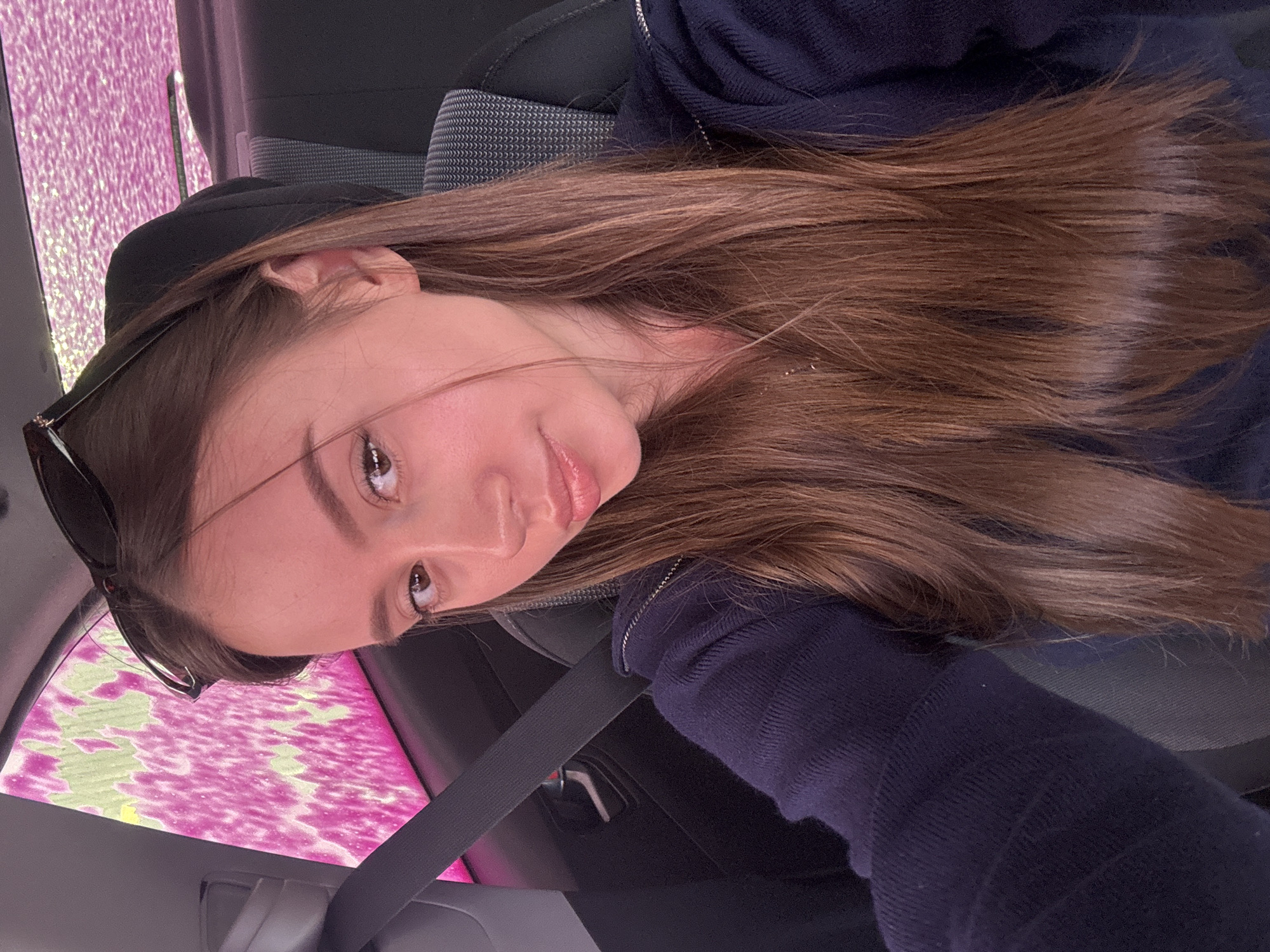A young woman is seated in a car, likely in the driver's seat, as indicated by the visible seatbelt across her chest. She has long, light brown hair and is wearing a dark blue zippered sweatshirt and sunglasses perched atop her head. The car window is showing a view of what appears to be a car wash, with pink and white soapy foam visible. The car seat is dark gray with a light gray pattern. The image is rotated, with the woman's face tilted towards the upper left. The city of Unknown, Ukraine, is not discernible in this image.