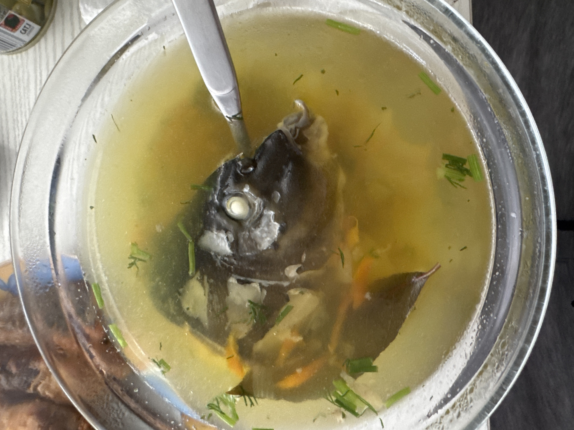 The image shows a close-up of a clear bowl of fish soup, possibly a bouillon, containing a fish head, some chopped green herbs, and what appear to be pieces of root vegetables like carrots and potatoes. A silver spoon is partially submerged in the soup, resting near the fish head. The soup is a golden-yellow color and appears to be hot, with some condensation visible on the rim of the bowl. The background is out of focus but suggests a table setting with some bread or pastry to the left and a jar with a label that reads "CAT.1" to the upper right. There is no identifiable location in the city of Duren, Germany visible in this image.