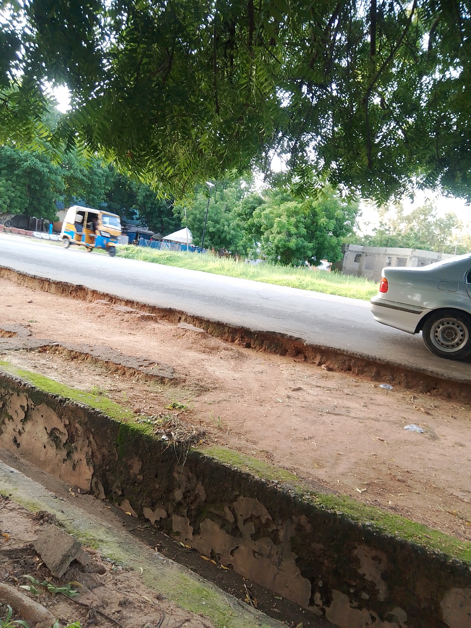 This media file captures an outdoor scene in Jalingo, Nigeria, likely during the daytime given the lighting. The foreground features a weathered, moss-covered concrete structure, possibly a drain or a raised planter, with eroded sandy soil alongside it. A paved road runs through the middle ground.
On the road, a yellow and blue motorized rickshaw, commonly known as an auto-rickshaw or "Keke Napep," is in motion, indicated by motion blur. Further down the road, the rear of a silver sedan is visible, parked or moving slowly. The background is filled with lush green trees and foliage, suggesting a well-vegetated area. Beyond the trees, a simple building structure with windows can be seen. In the distance, a white tent-like structure is also present.
The overall impression is a snapshot of everyday life in a tropical or subtropical urban environment. The presence of a Keke Napep points to local transportation. The eroded roadside suggests potential issues with infrastructure or natural elements like rainfall. The clear visibility and the foliage indicate a dry or moderate weather condition. There is no visible text in the image.