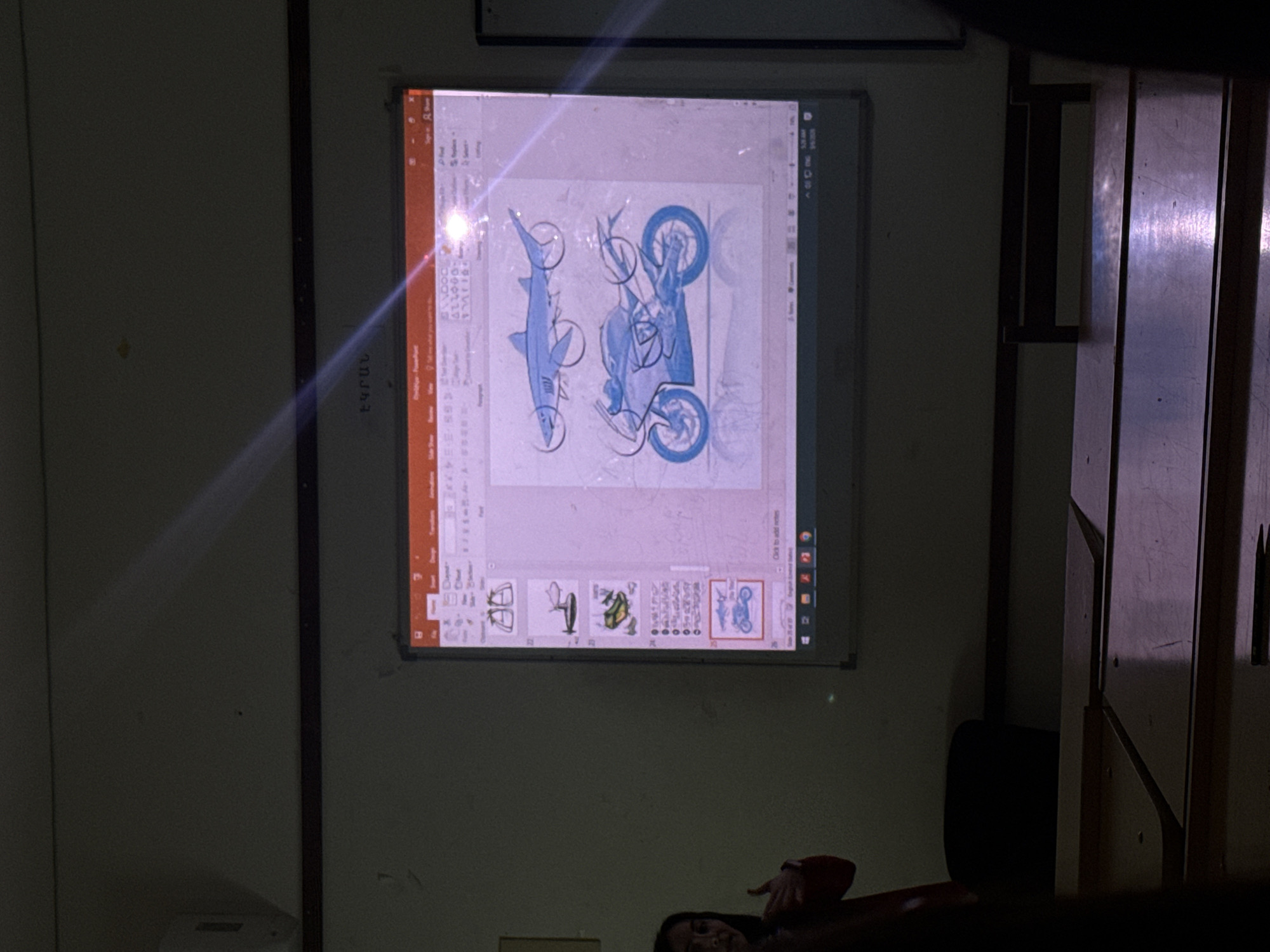 A projected image from a smartboard in a classroom shows a Microsoft PowerPoint presentation. The presentation has several slides previewed at the bottom, including images of a shark, a motorcycle, and other objects. A person is partially visible at the bottom edge of the frame. The classroom has light-colored walls and a combination of dark and light furniture. The image does not provide information to determine the location in the city of Yerevan, Armenia.