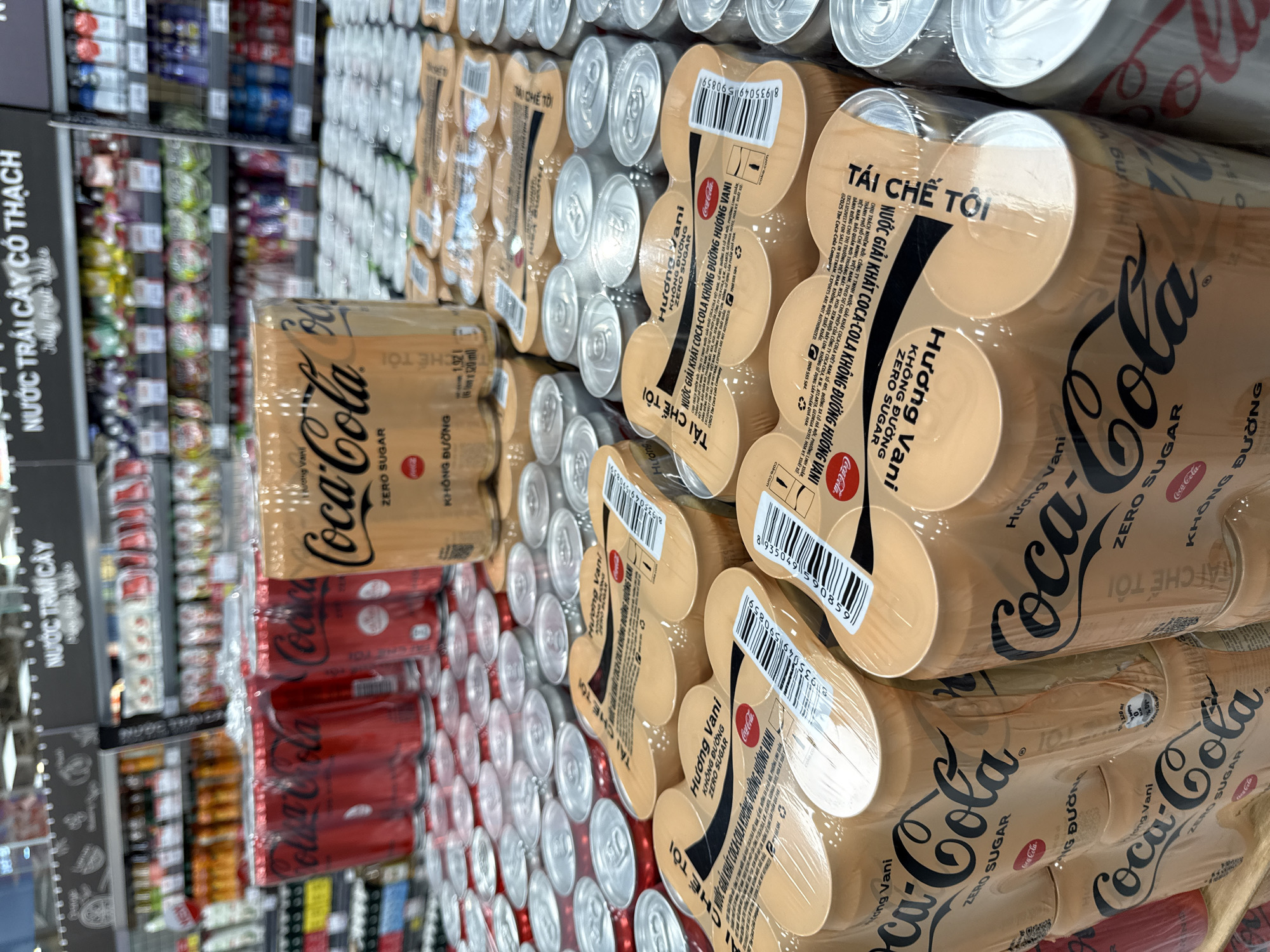 In a retail setting, likely a supermarket aisle in Nha Trang, Vietnam, shelves are stocked with beverages. The foreground is dominated by multiple packs of Coca-Cola Zero Sugar cans, specifically in a vanilla flavor indicated by "HƯƠNG VANI KHÔNG ĐƯỜNG ZERO SUGAR" printed on the packaging. These packs contain six cans each, wrapped in clear plastic. The cans themselves are primarily gold with the iconic Coca-Cola logo in black. Behind these packs, a variety of other canned drinks are visible, including red Coca-Cola cans and silver-colored cans. The background reveals more shelves filled with assorted packaged drinks and products, suggesting a well-stocked store. The lighting appears to be artificial, typical of an indoor retail environment. There are no people or discernible actions in the immediate foreground or midground.
