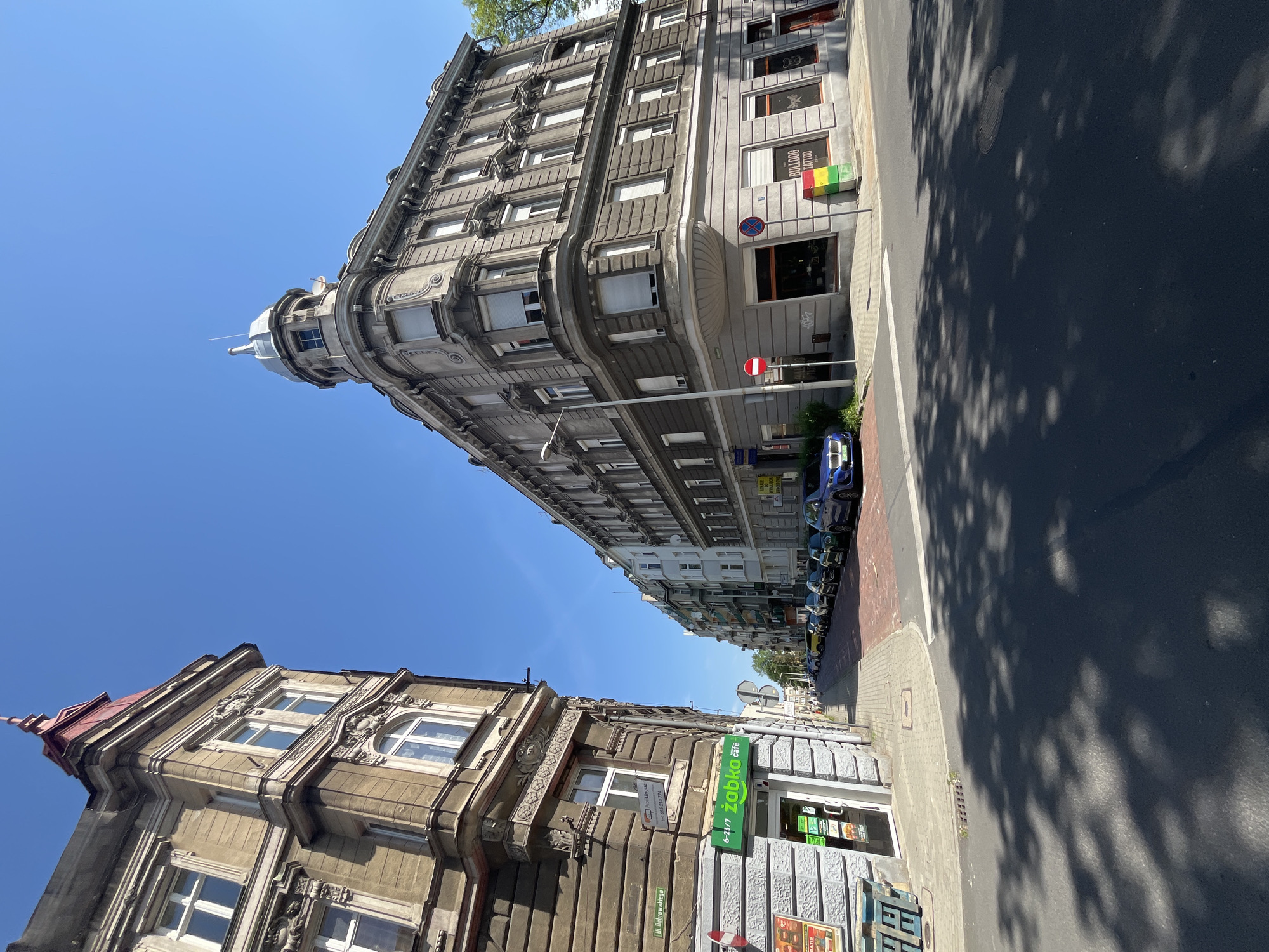 The image shows a street intersection in a city, likely in Europe based on the architecture and street signs. On the left is a multi-story building with ornate detailing and a red-roofed spire. Adjacent to it, at the corner, is a shop with a green "Żabka" logo, which appears to be a convenience store. Across the intersection, on the right, is another large, ornate building with a domed turret. This building houses a tattoo parlor called "BULLDOG TATTOO". Several cars are parked along the street in the mid-ground. The street is paved with asphalt, and there are trees casting shadows on the road, indicating it is daytime. The sky is clear and blue. The general impression is of an older, well-established urban area.