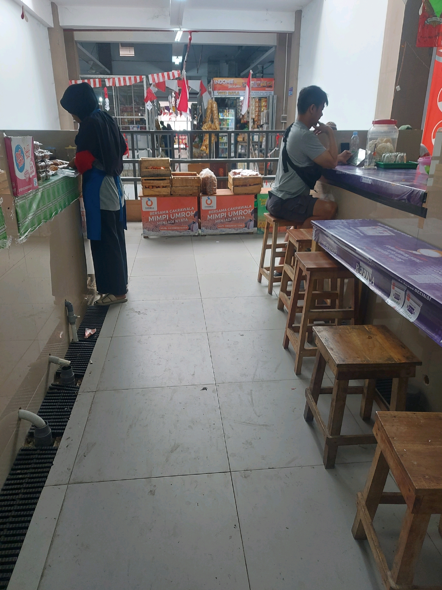 The image captures a scene inside a food stall or small eatery, likely located in Godean, Indonesia, judging by the context of the surrounding businesses. A woman wearing a black hijab, a red top, and a blue apron stands behind a counter, appearing to prepare or serve food. In front of her, there are stacks of what appear to be food products in boxes and crates.
To the right of the image, a man is seated on a stool at a counter, looking down at his phone. He is wearing a grey t-shirt and dark shorts. The counter itself is covered with a purple tarp-like material, and there are various items on it, including a clear plastic jar filled with crackers or snacks, a bottle of water, and a small plate with green items. Several wooden stools are lined up along the counter, some occupied and some empty.
The environment appears to be somewhat utilitarian, with a tiled floor that shows signs of wear and dirt. A metal grate runs along the wall near the counter, with some plumbing visible underneath. In the background, shelves stocked with products and more promotional materials are visible, suggesting a bustling commercial space. Decorative red and white banners are hung across the ceiling, hinting at a festive occasion or holiday. The overall impression is of a local, informal food service establishment.