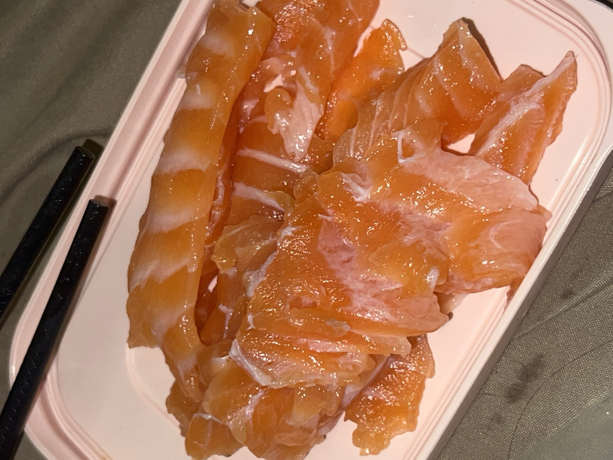 The image presents a high-angle, close-up view of numerous pieces of thinly sliced, raw orange-pink fish, most likely salmon, arranged on a pale pink, rectangular tray. The fish pieces vary in shape and size, displaying a glossy, slightly translucent appearance with distinct white marbling or streaks throughout, suggesting freshness.
The tray has a simple design with a raised rim and is positioned on a dark, textured fabric surface, which appears to be olive green or dark grey. To the left side of the tray, two dark, slender objects resembling chopsticks are partially visible, resting on the fabric.
The overall lighting is somewhat dim, creating soft reflections on the surface of the fish, which points to an indoor setting. The presence of chopsticks alongside the raw fish suggests the dish is intended for an Asian meal, possibly sashimi or a component for a hot pot. No specific time of day, weather conditions, people, or discernible emotions are evident in the image. There is no visible text. The scene depicts food prepared for consumption. The context provided indicates this event took place in Bac Nha Trang Ward, Vietnam.