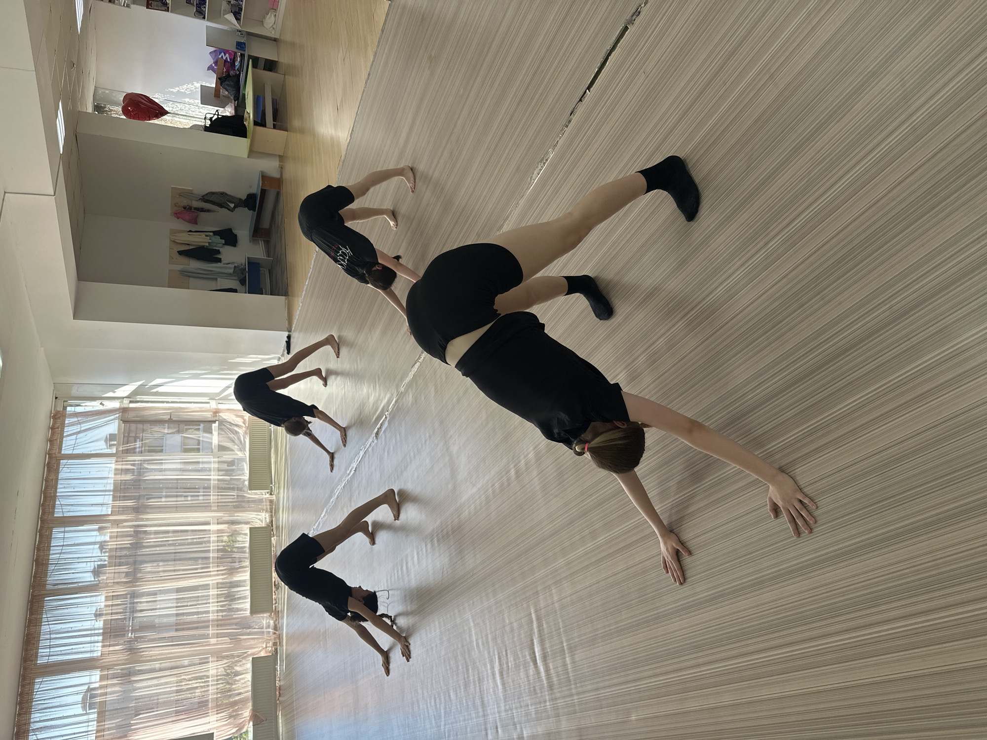 Four people are practicing yoga in what appears to be a dance or yoga studio. They are performing a downward dog pose. The studio has light-colored wooden floors and large windows covered with sheer curtains. The location appears to be in Votkinsk, Russia, although this cannot be verified from the image alone. Shelves are visible at the back of the room.
