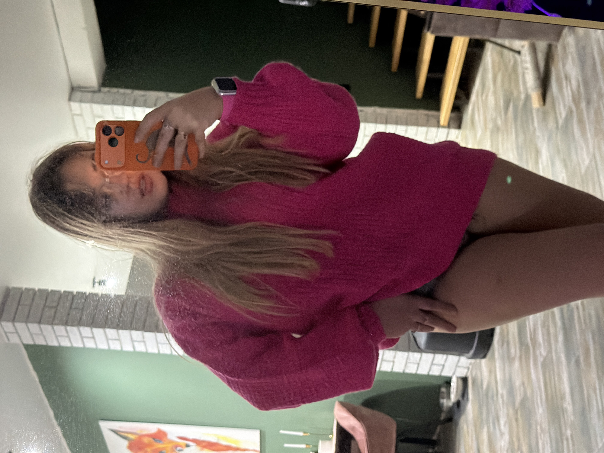 A woman is taking a selfie in a mirror. She is wearing a pink sweater. The background includes a green wall, a painting, and a staircase. The location is near Большая Шильна, Russia.