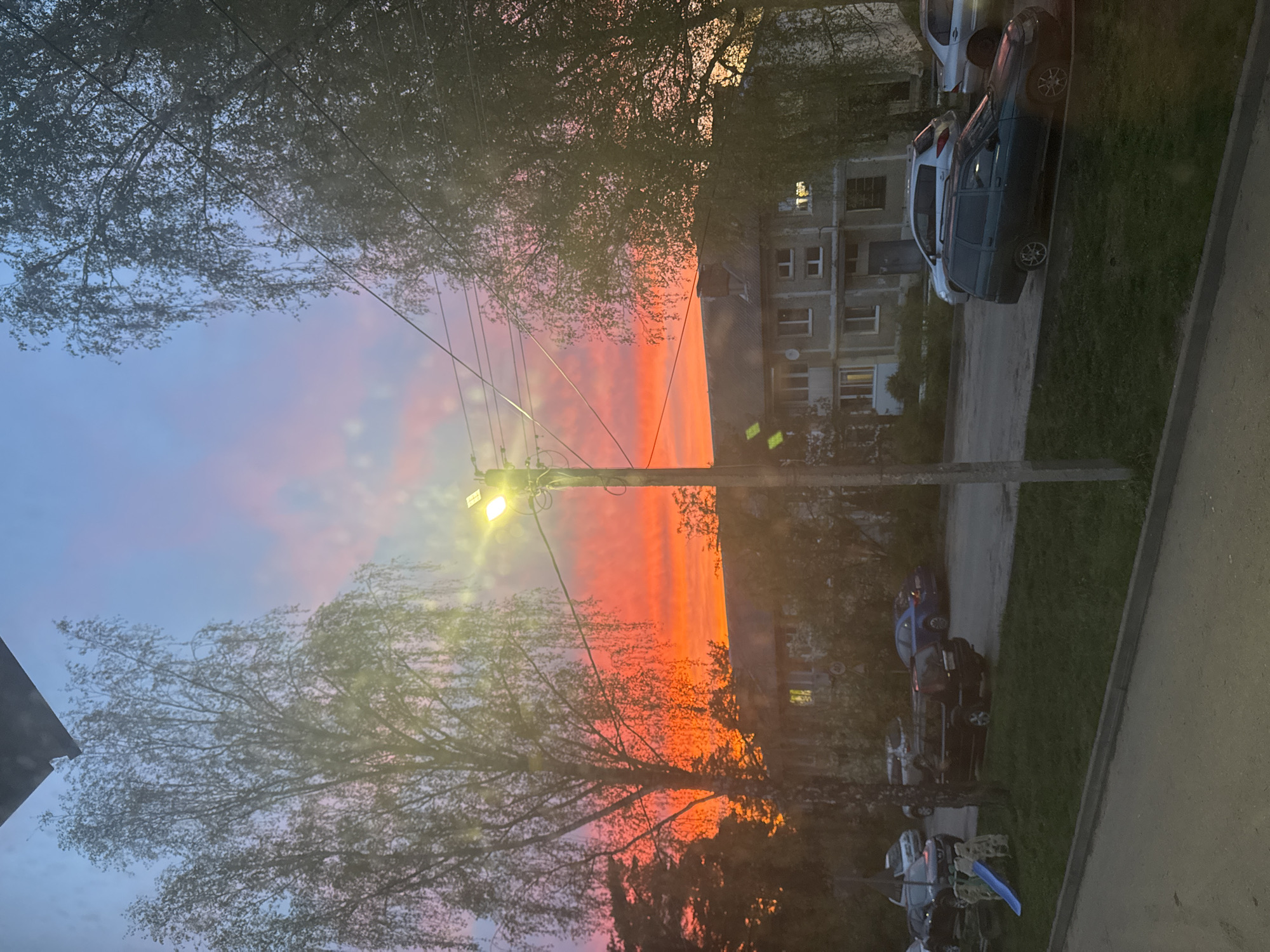 The image depicts a vivid sunset sky with red and orange hues visible through the branches of trees. A utility pole with a street light is prominent in the center. In the background, there is a multi-story residential building, and several cars are parked along a roadway. The scene appears to be in a residential area in Раздолье, Russia, based on the location context provided. The image seems to have been taken from an elevated position, possibly from a window.