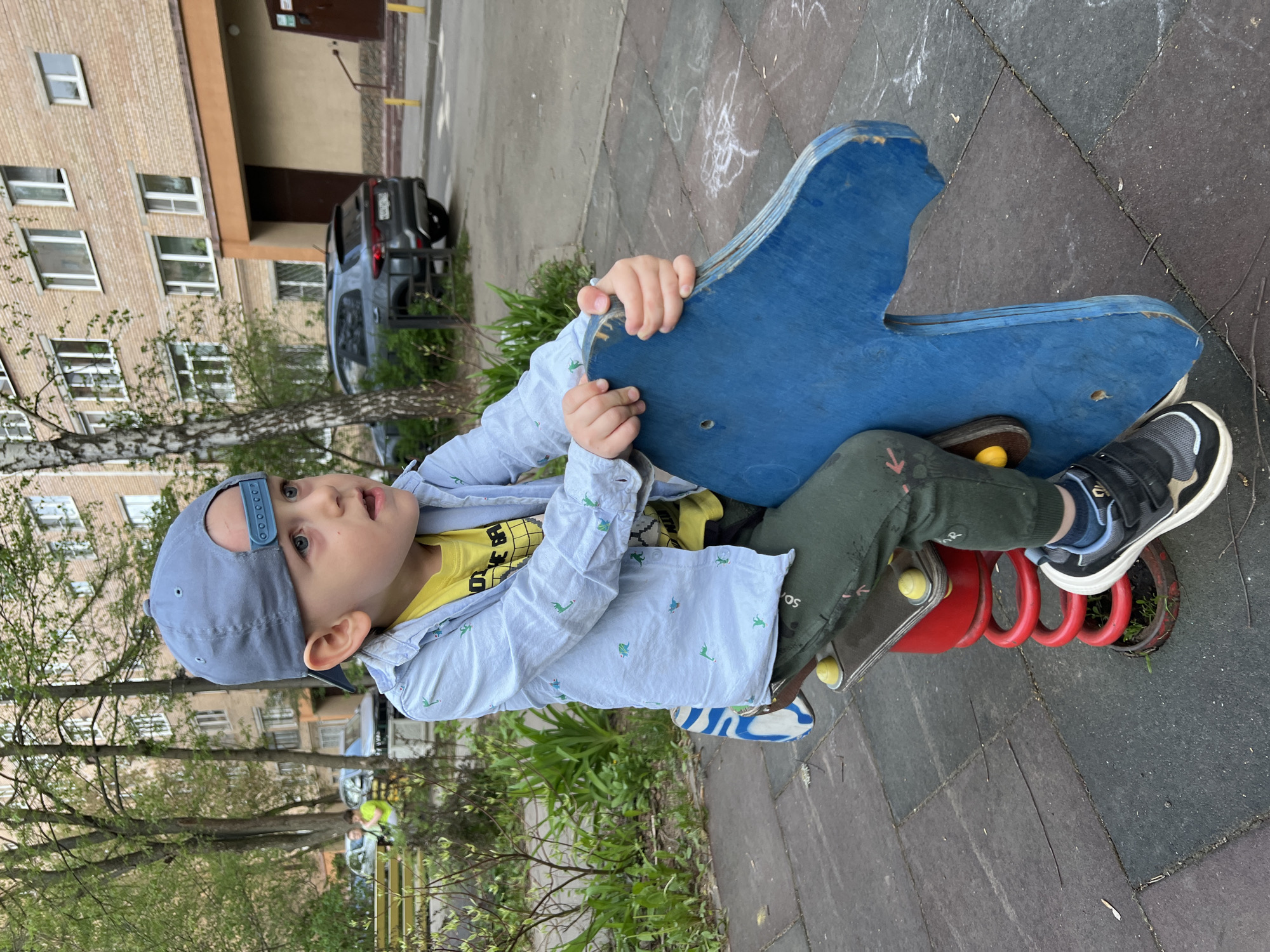 A young boy is sitting on a blue, wooden, spring-mounted rocking horse in a playground. He is wearing a light blue shirt with a pattern of dinosaurs, a yellow t-shirt, green pants, a gray baseball cap, and black and white sneakers. The rocking horse has a red spring base. The playground is located near a building with multiple windows and a parked car. Based on the architectural style and vegetation, the location is consistent with Mytishchi, Russia.