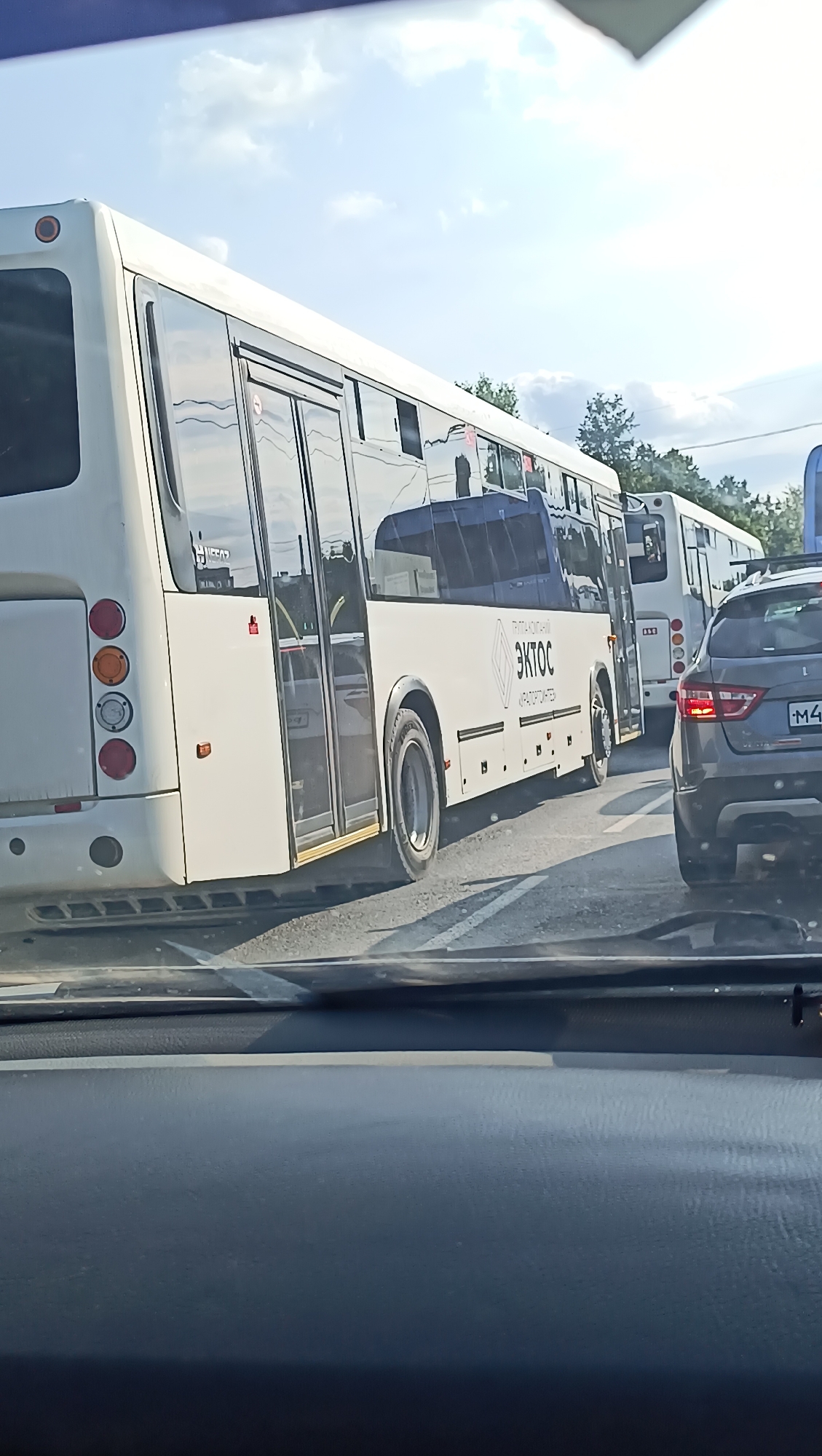 The image shows a line of buses and a car on a street, likely in the city of Chaikovsky, Russia. The buses are white, with one prominently displaying the logo "ЭКТОС" (EKTOS) and text below it that appears to be in Russian. The car in the foreground is dark gray. The sky is visible, with some clouds and trees in the background. The scene appears to be a traffic jam.