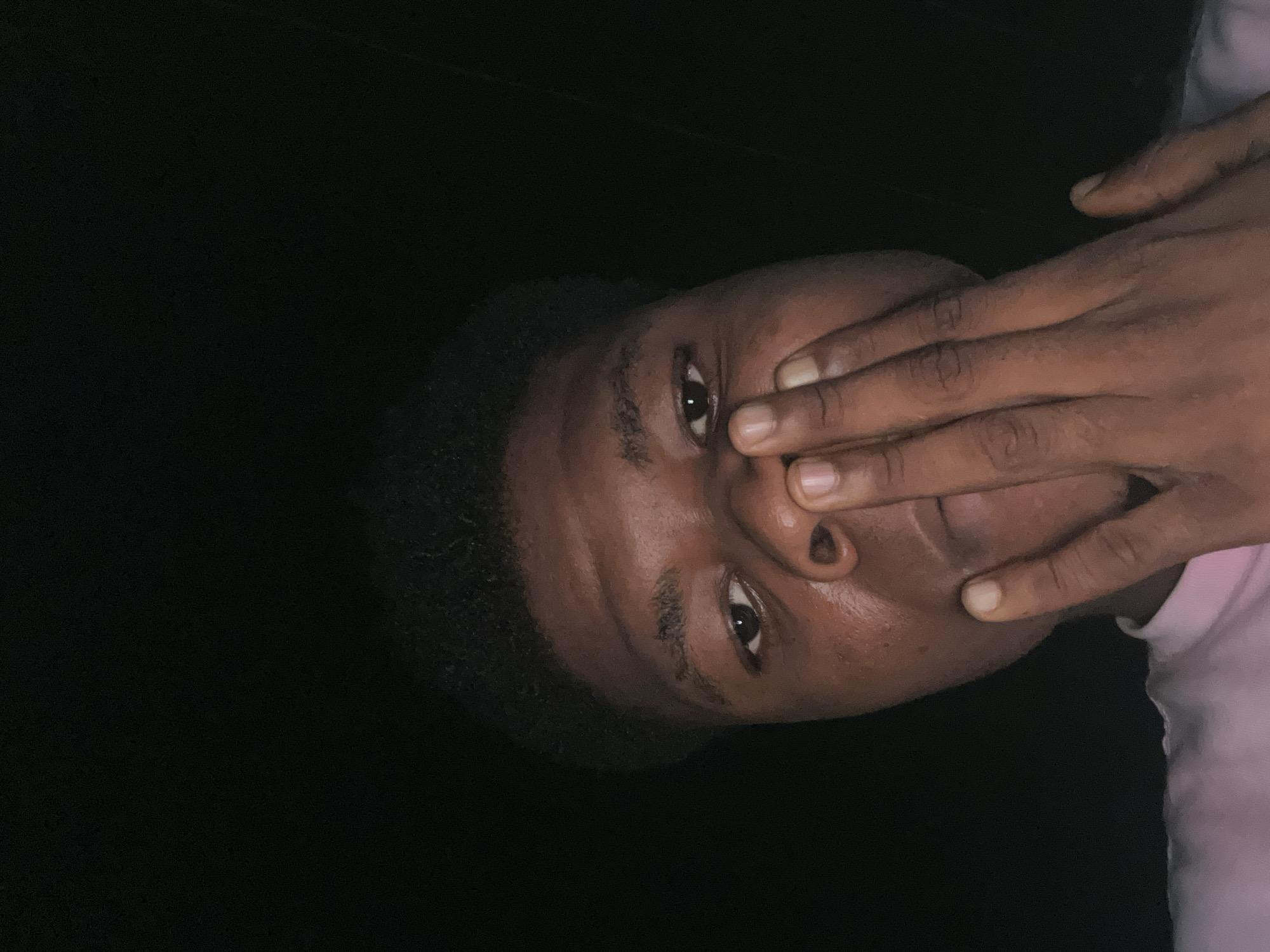 The image is a close-up selfie of a young Black person's face, partially obscured by their hand. The person has dark, short, curly hair and is looking directly at the camera with their right eye visible. Their left hand is positioned over the lower part of their face, with fingers covering their mouth and nose. The lighting is dim, casting shadows and highlighting the contours of their face. The background is entirely black, suggesting a dark environment, possibly at night. The person is wearing a pink t-shirt. The image appears to have been taken indoors or in a place with minimal ambient light, and is likely a self-portrait taken by the subject themselves. There is no visible indication of the location being within the city of Ihiagwa, Nigeria.