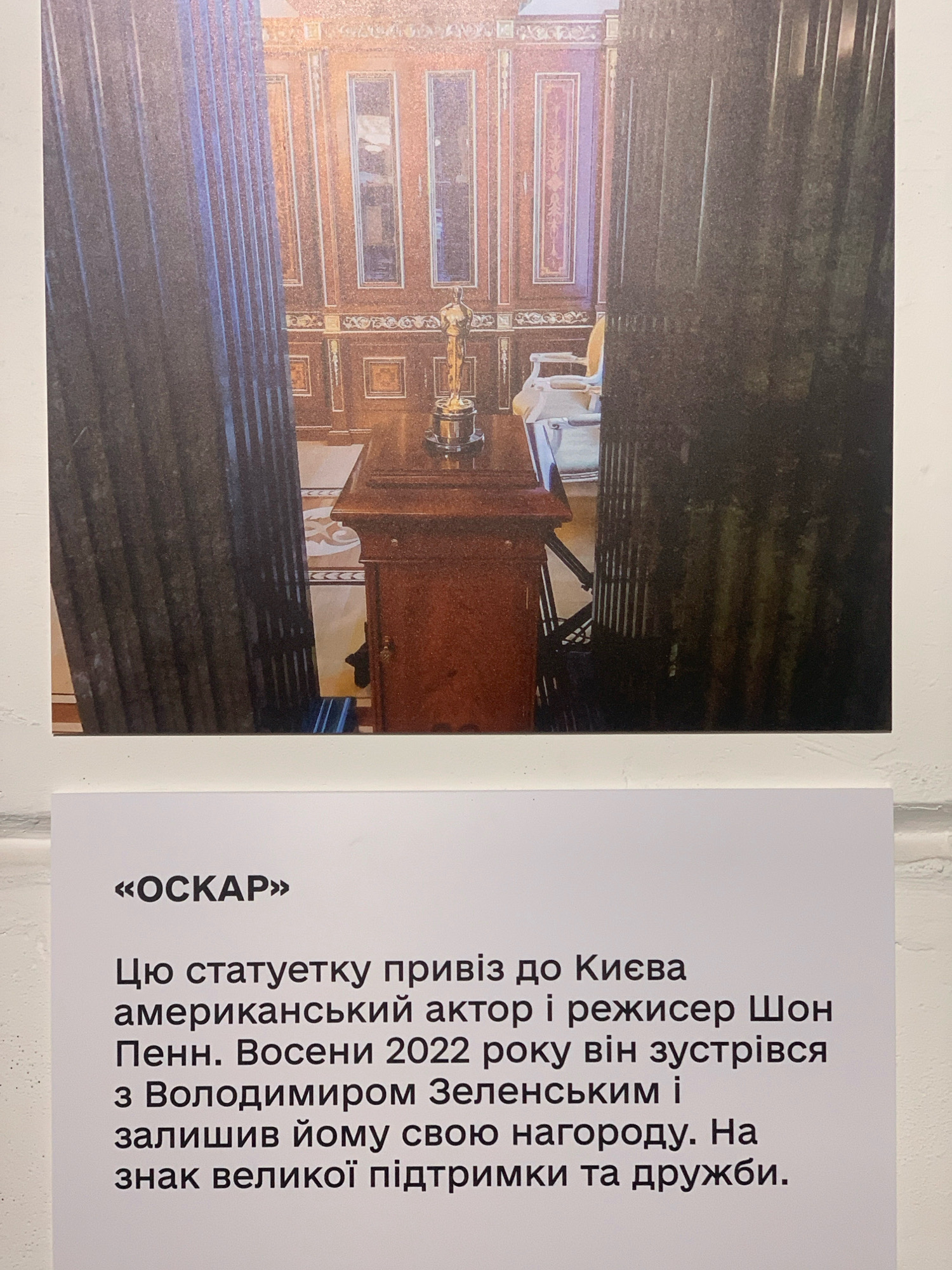 The image displays a photograph of an Oscar statue placed on a wooden pedestal in an ornate room, likely a government building, with large columns framing the view. Below the photograph, a white sign contains text in Ukrainian. The text translates to: "OSCAR. This statue was brought to Kyiv by the American actor and director Sean Penn. In the autumn of 2022, he met with Volodymyr Zelensky and left him his award as a sign of great support and friendship." While the exact location within Kyiv is not specified, the ornate interior and the presence of government officials suggest it is likely a formal setting such as the Presidential Office or a similar state institution. The photograph itself focuses on the Oscar statue and the rich, classical interior.