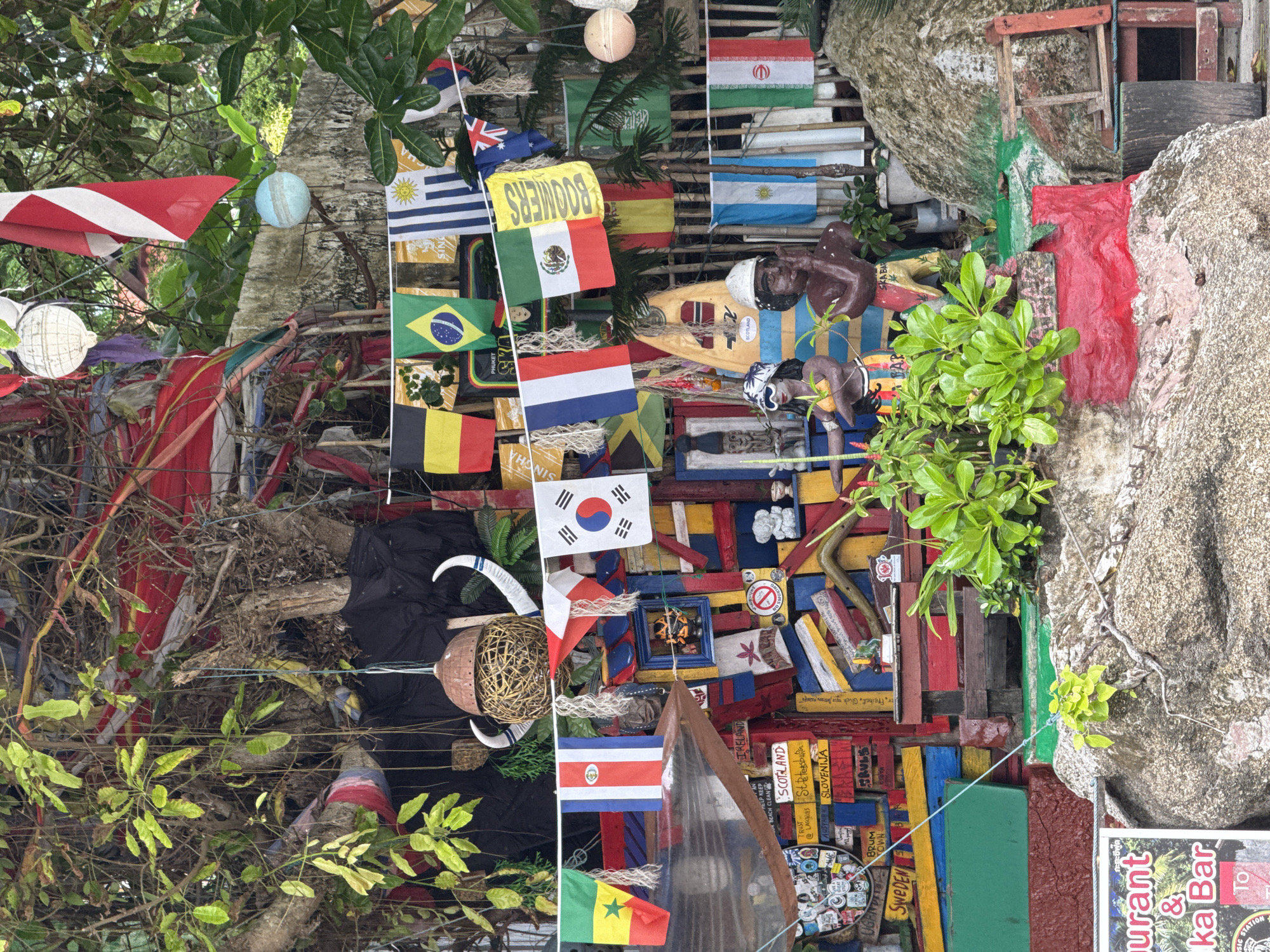 This image depicts a colorful and eclectic display of international flags, souvenirs, and decorative objects arranged vertically against a natural background of tree branches and a rocky surface. The flags of many nations, including the USA, Mexico, Brazil, South Korea, Spain, and Iran, are prominently displayed, suggesting a global theme or a collection of travel memorabilia. Interspersed among the flags are various trinkets, small sculptures of people, and what appear to be handmade decorations. A sign that reads "BOOMERS" is visible, as is a sign for "SKA BAR" and "Karon, Thailand". The scene is bustling with visual information, with layers of objects and textures creating a sense of abundance and perhaps a vibrant marketplace or a personal collection displayed outdoors. The lighting suggests it is daytime, with natural light illuminating the scene. The overall impression is one of a lively, international, and perhaps tourist-oriented location.