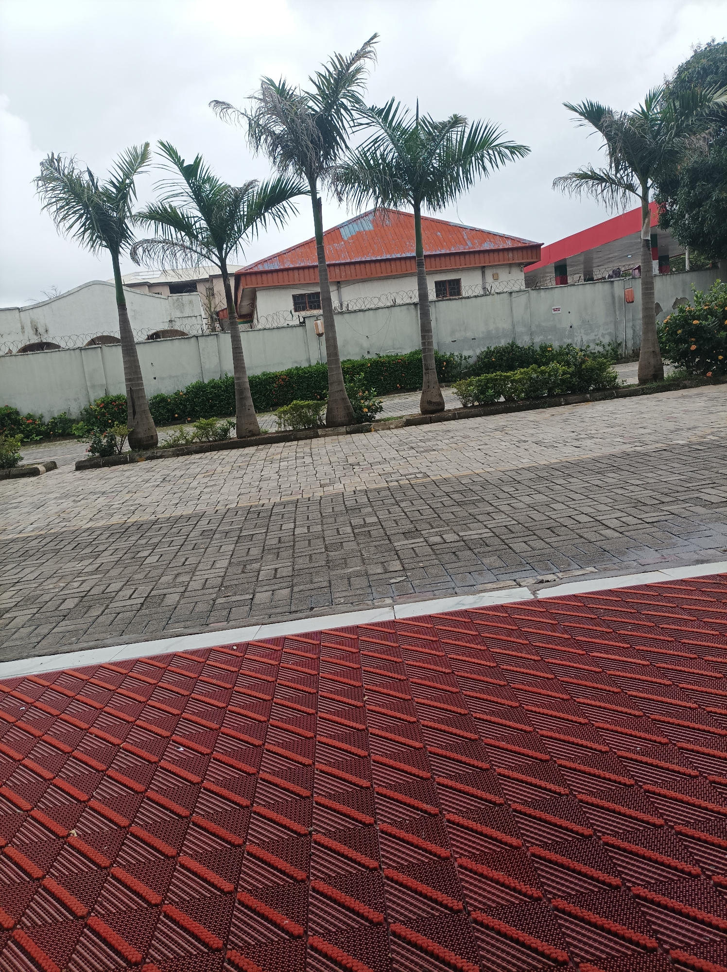 The image captures an outdoor scene with a paved area in the foreground and a building with palm trees in the background. The foreground consists of a red, textured surface with a patterned design, transitioning to a gray, brick-like paving. In the middle ground, a row of palm trees stands in front of a wall. Behind the wall, a building with a reddish-orange roof is visible. The sky is overcast. The location is identified as Abuja, Nigeria.