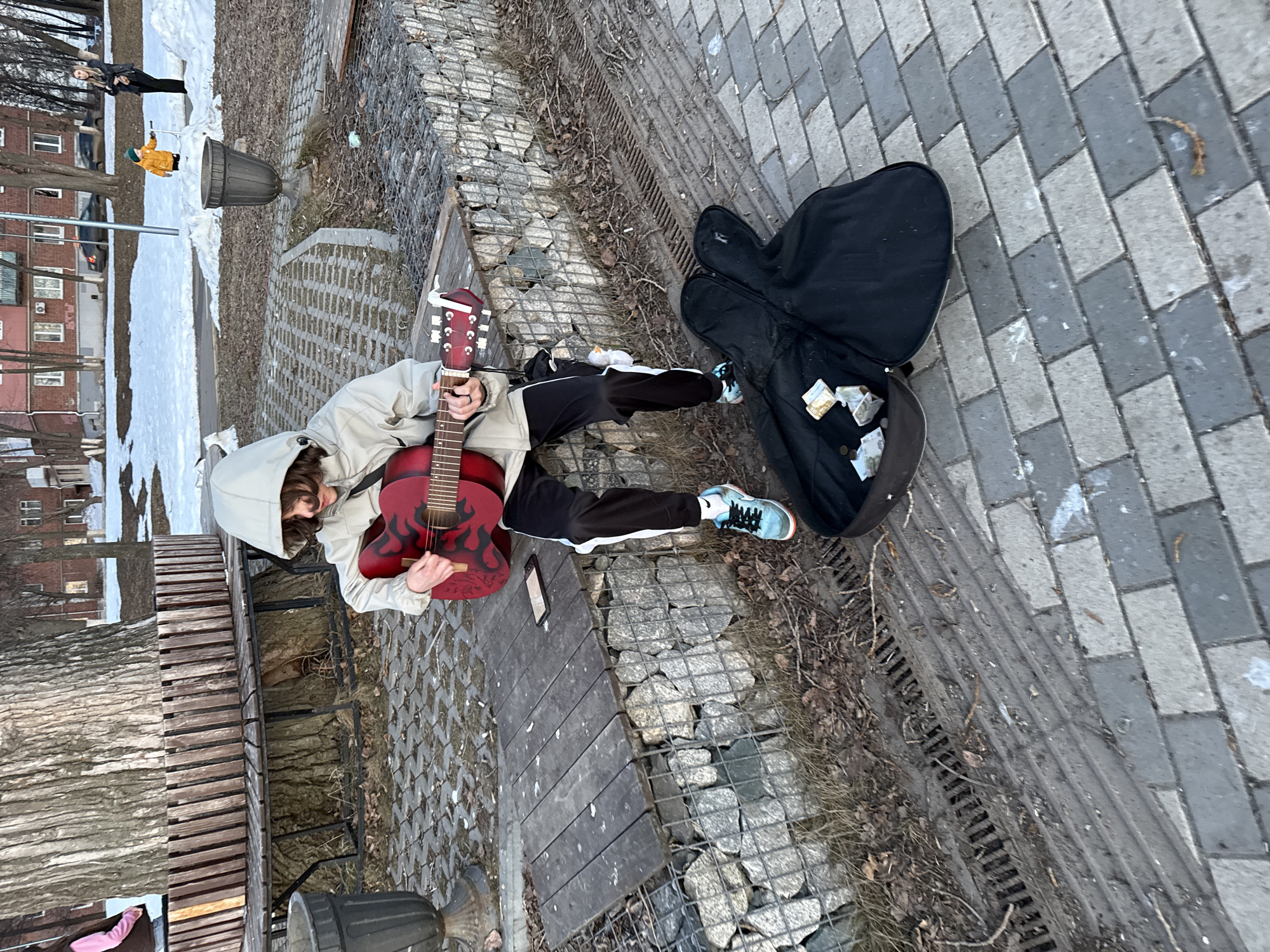 A person with a hooded jacket is sitting on a bench in an outdoor urban setting, playing a red electric guitar. The person is wearing black and white striped pants and light blue sneakers. Next to them is an open guitar case with money inside. The background includes snow-covered areas, buildings, trees, and other people walking. The setting appears to be a park or public space in Votkinsk, Russia. The ground is partially paved and has debris and leaves scattered around.